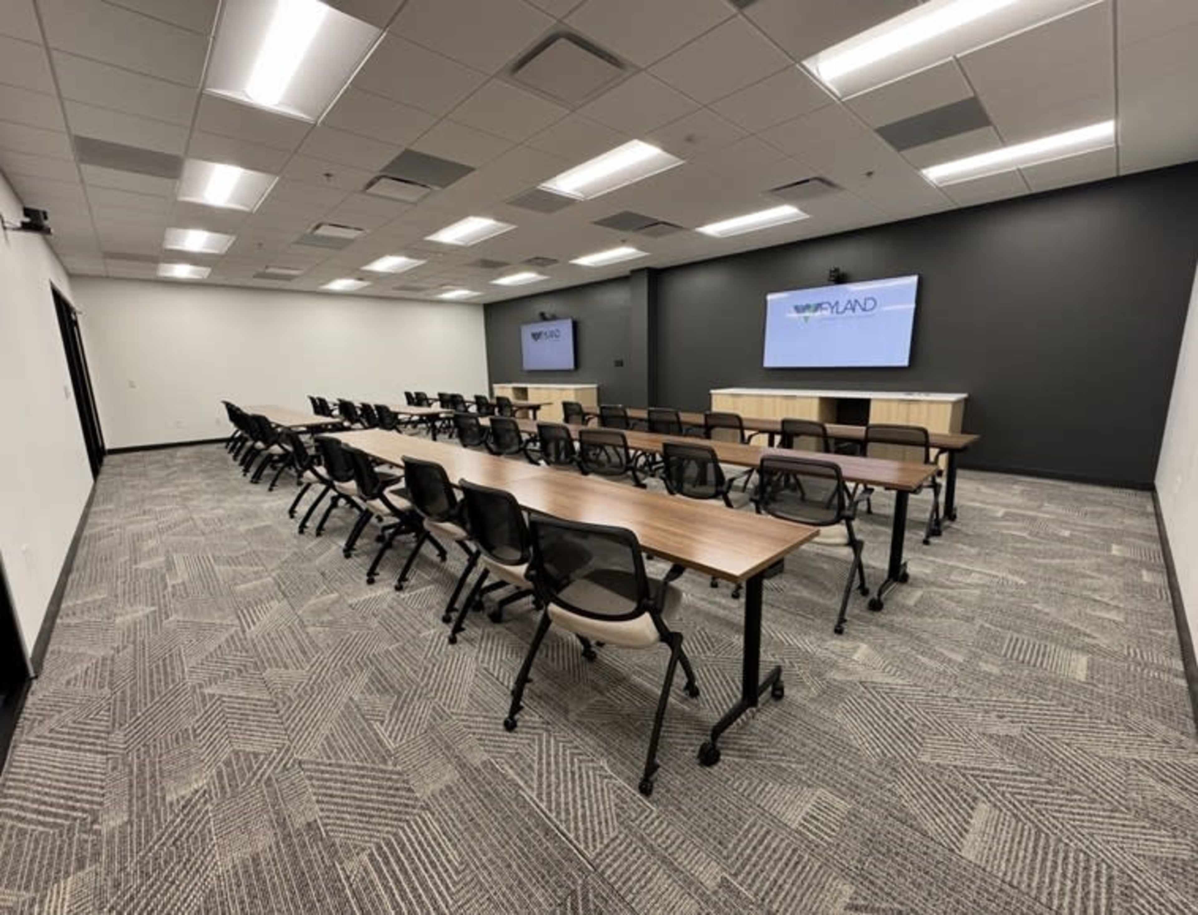 The image shows a modern conference room equipped with multiple tables and chairs arranged for a meeting, along with two large screens on the walls.