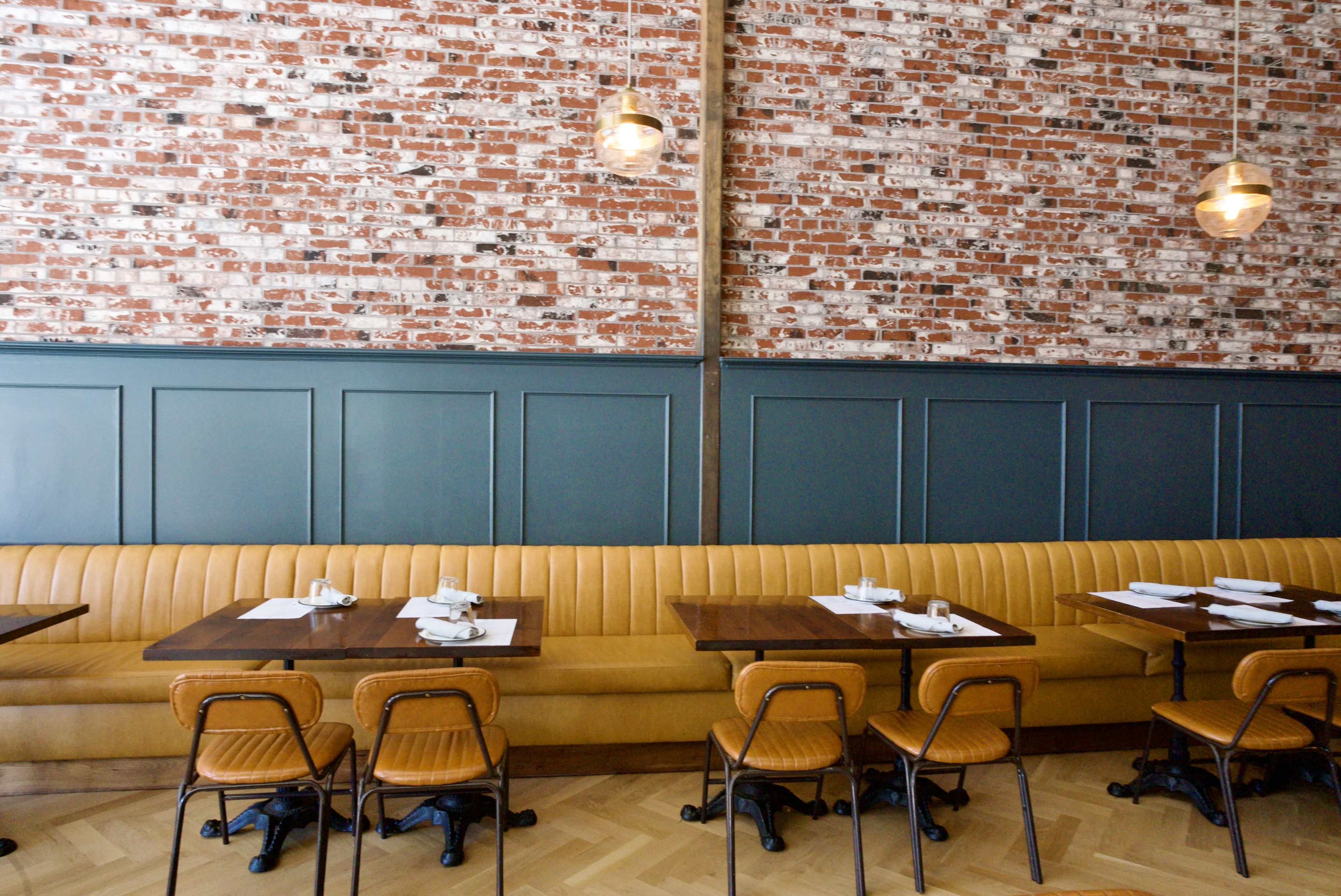 The image shows a modern restaurant interior with a brick wall, green paneling, and a long orange upholstered bench alongside several wooden tables and chairs.