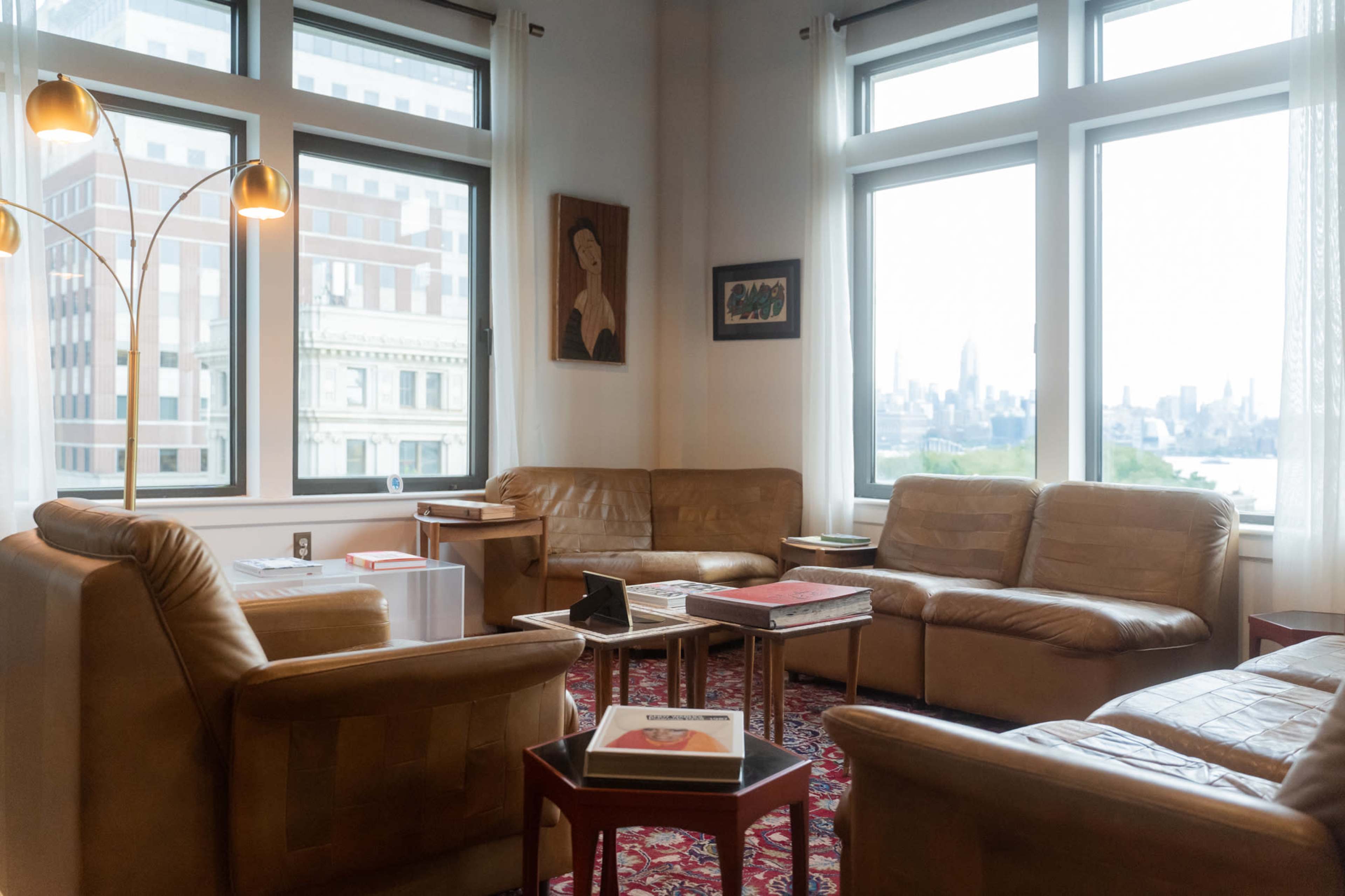 A cozy living room features two leather sofas, a modern floor lamp, a small table, and large windows overlooking a city skyline.