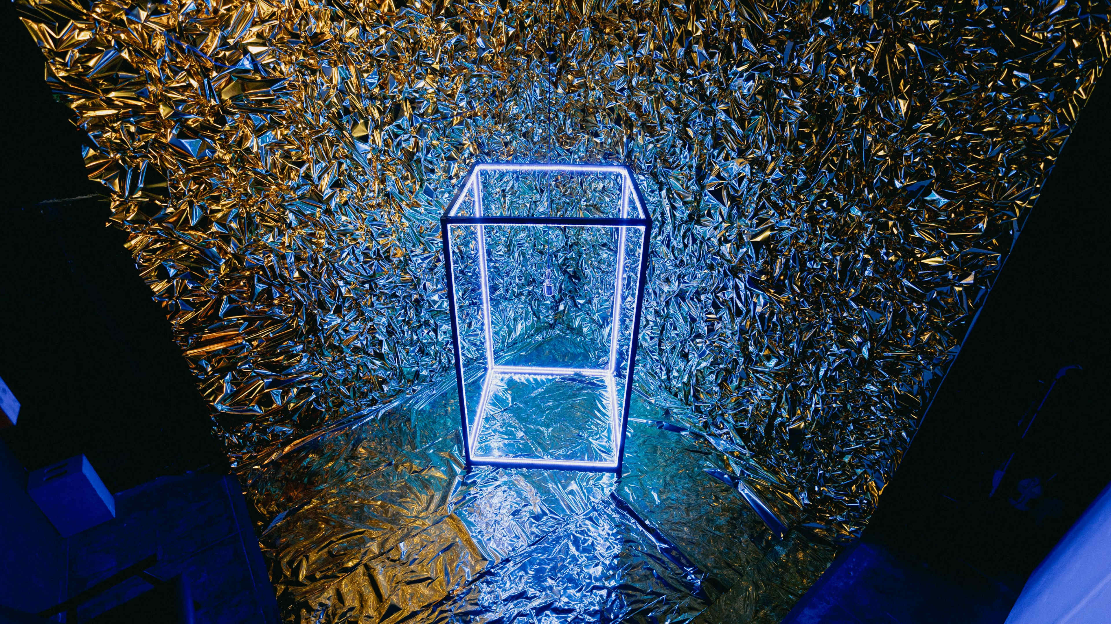 A transparent display box is illuminated by blue and white lights in front of a shiny gold and blue foil backdrop.