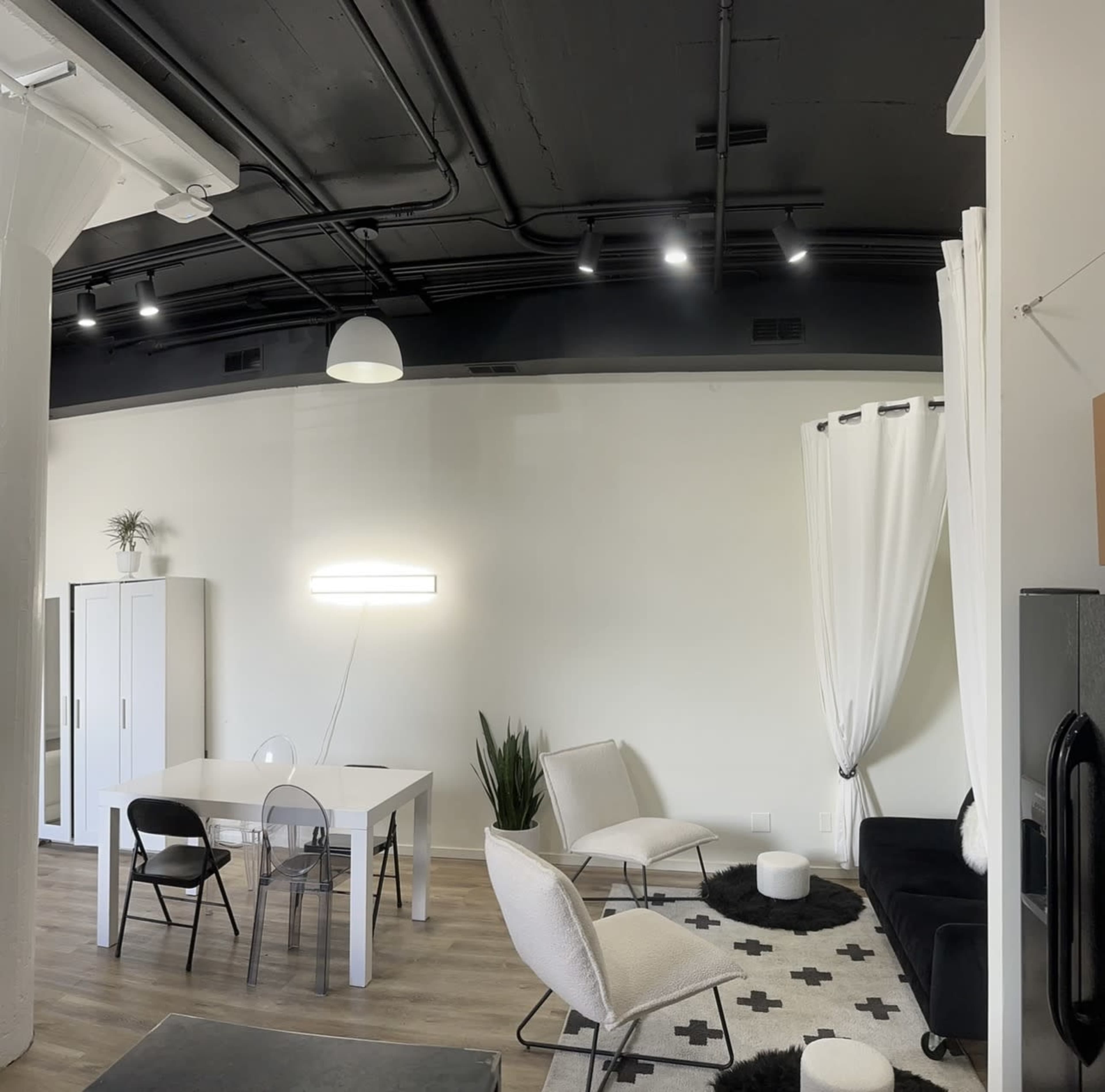 The image shows a modern, minimalistic living space with a white dining table, several chairs, and a black couch, complemented by indoor plants and a well-lit ceiling.