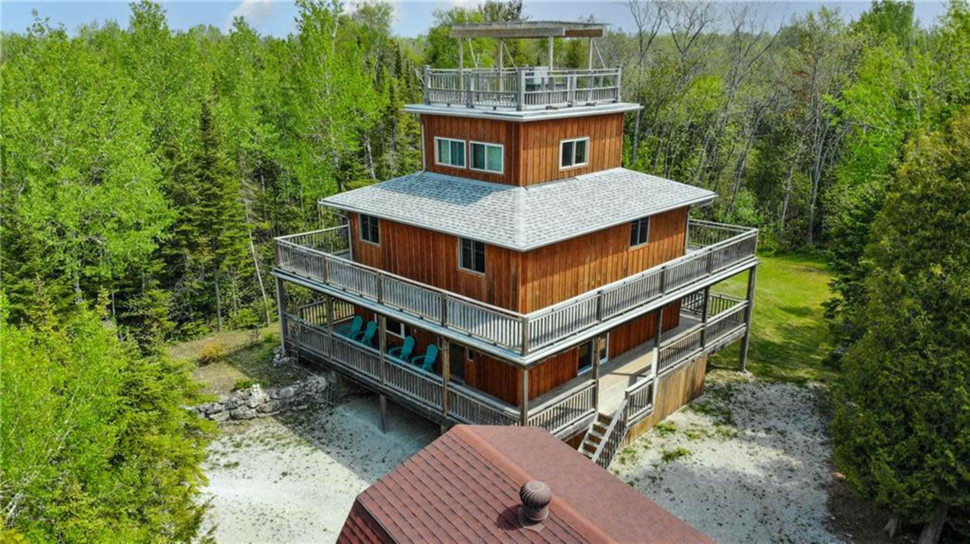 A modern, multi-level wooden cabin is surrounded by greenery, featuring multiple decks and a rooftop terrace.