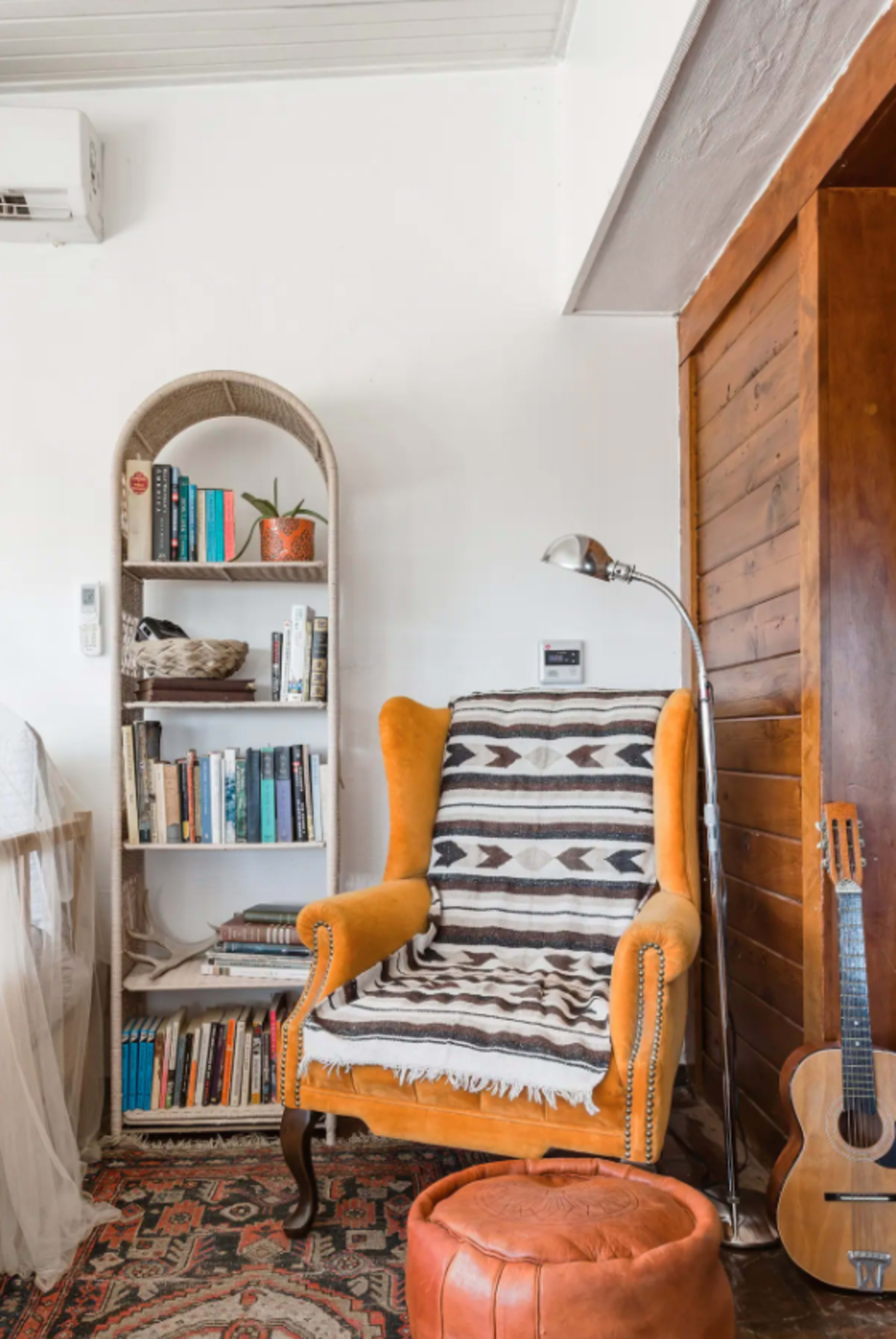 A cozy corner of a room featuring an orange wingback chair with a patterned throw, a bookshelf filled with books, a floor lamp, a leather pouf, and a guitar resting against the wall.