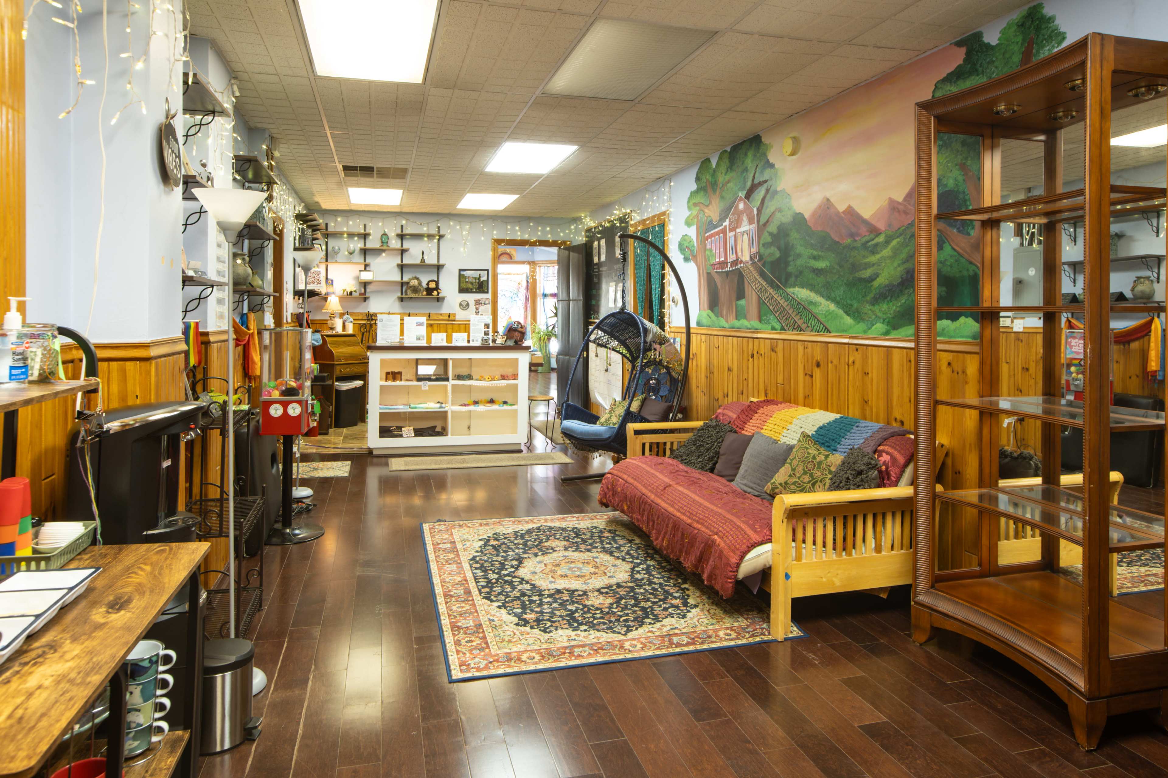 The image shows a cozy shop interior with wooden walls, a seating area featuring a patterned blanket, shelves displaying various items, and a mural of mountains in the background.