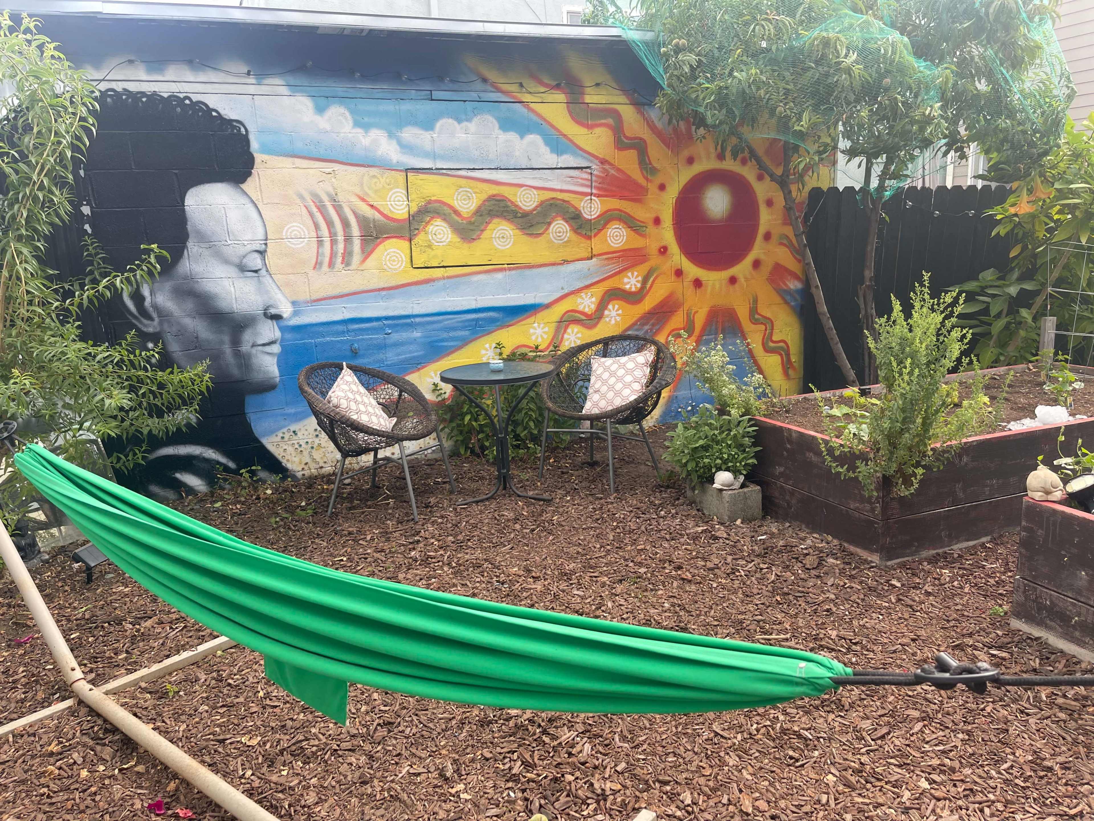 A green hammock is suspended in a garden featuring a mural of a person's profile beside a bright sun, with two chairs and a small table positioned nearby.