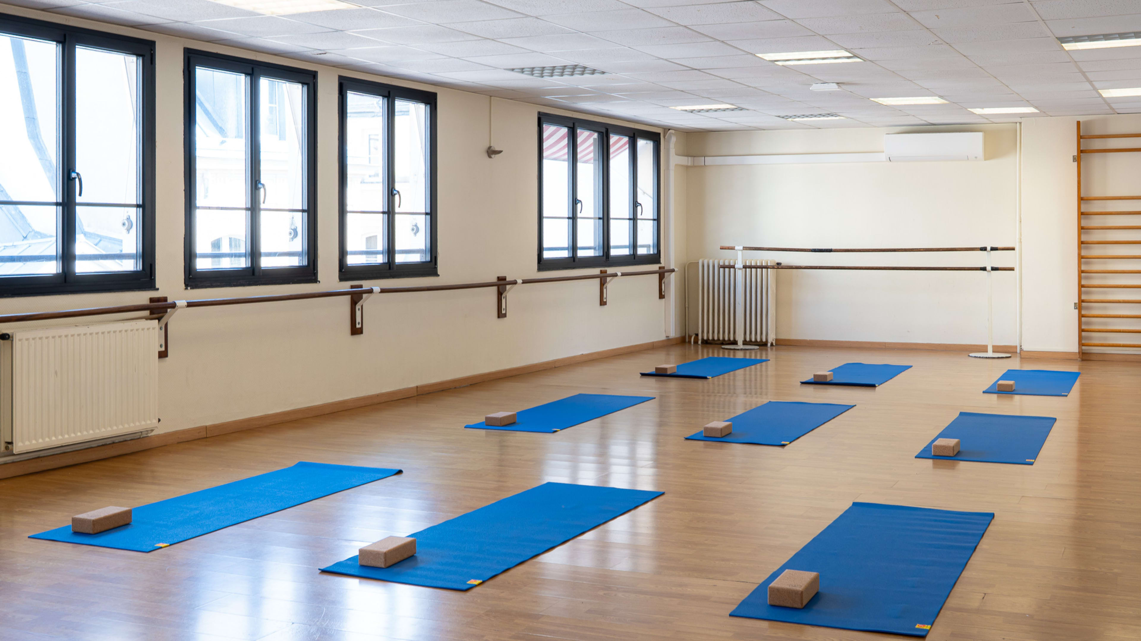 The image shows a spacious yoga studio with blue mats arranged on a wooden floor, each mat paired with a block, and large windows allowing natural light to enter.