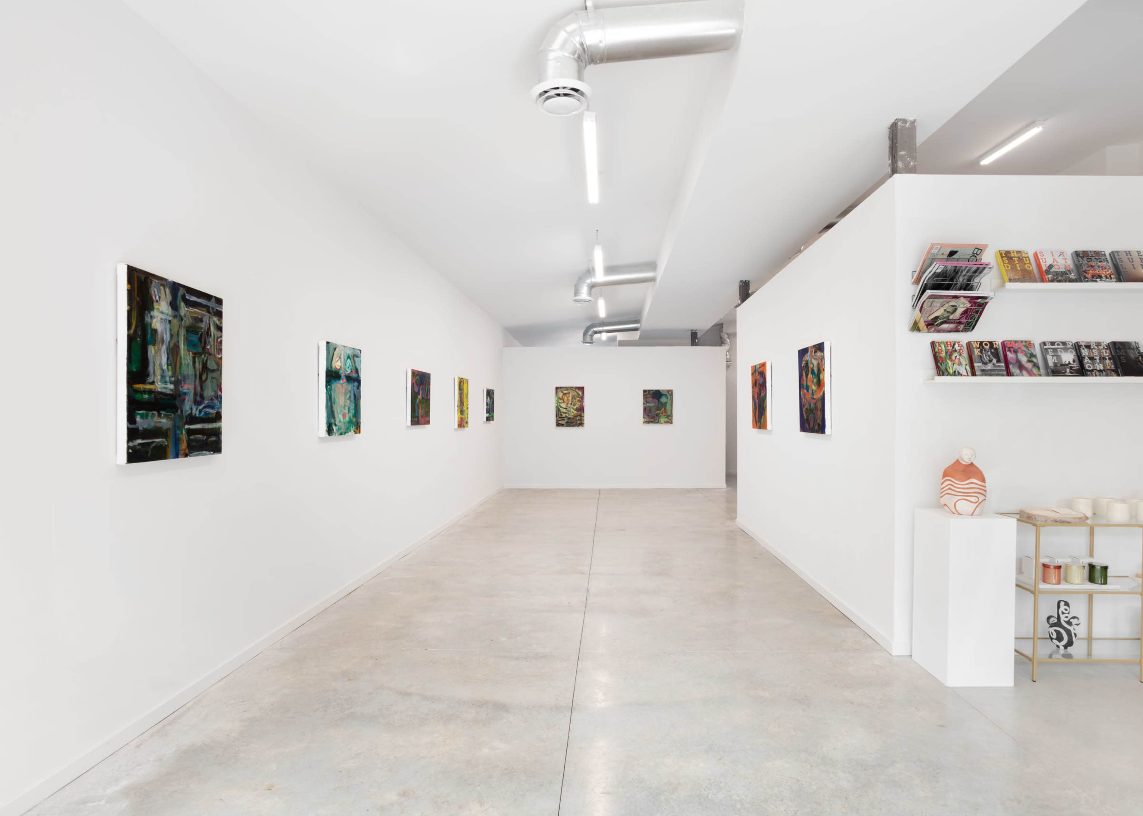 A minimalist art gallery featuring several framed paintings on white walls and a shelf displaying books and ceramics.