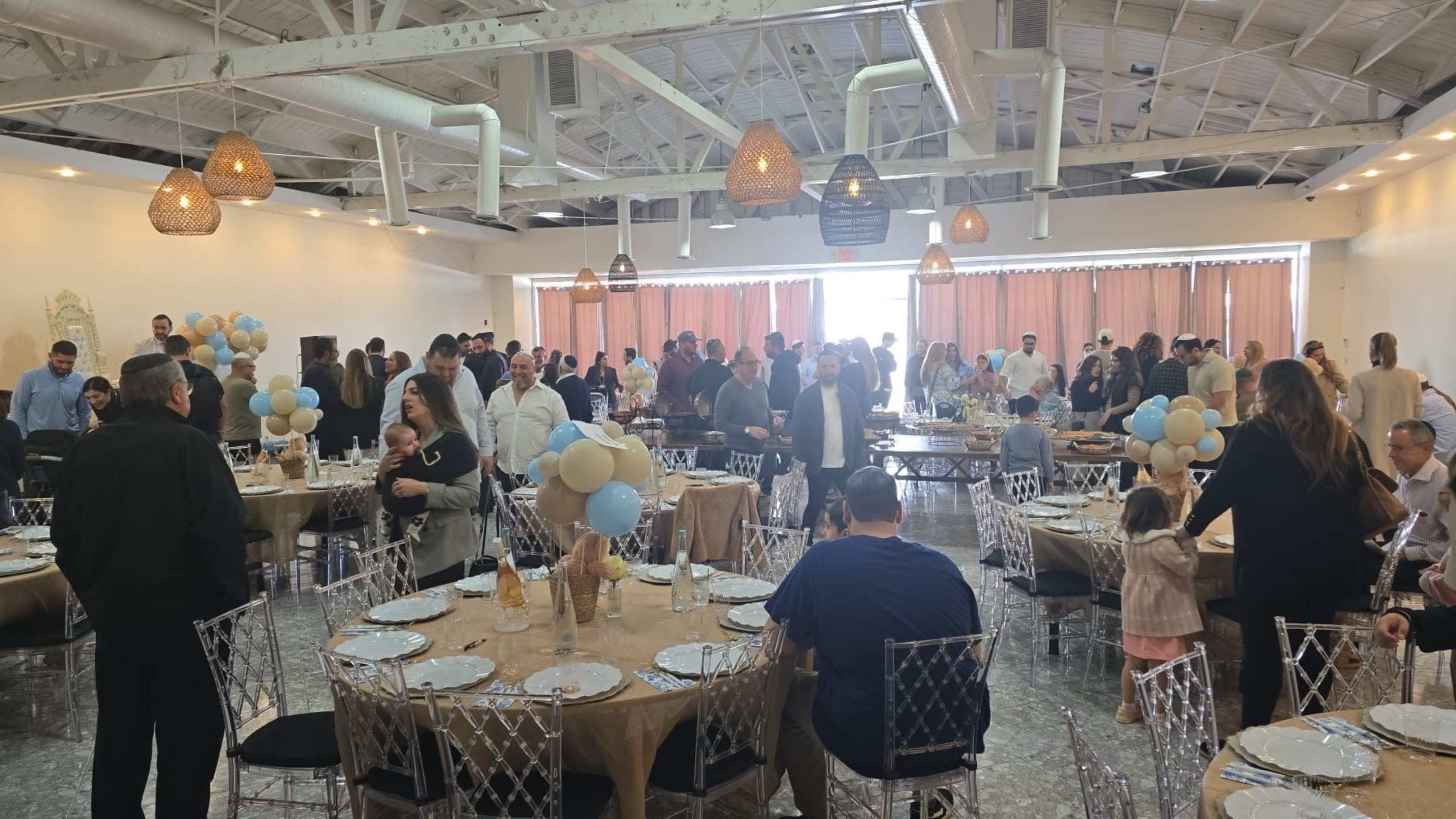 The image shows a large indoor event space filled with people mingling around elegantly set tables, decorated with balloons.