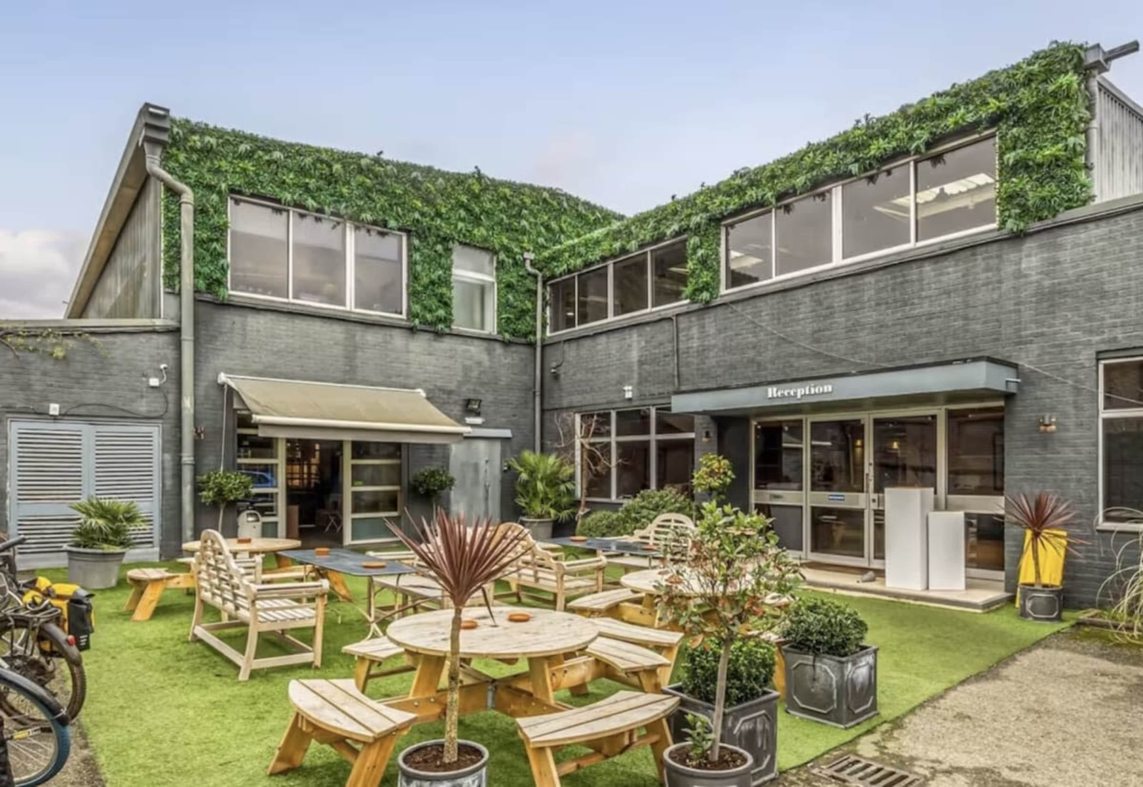 The image shows a modern building with a green roof, outdoor seating areas, and a reception entrance.