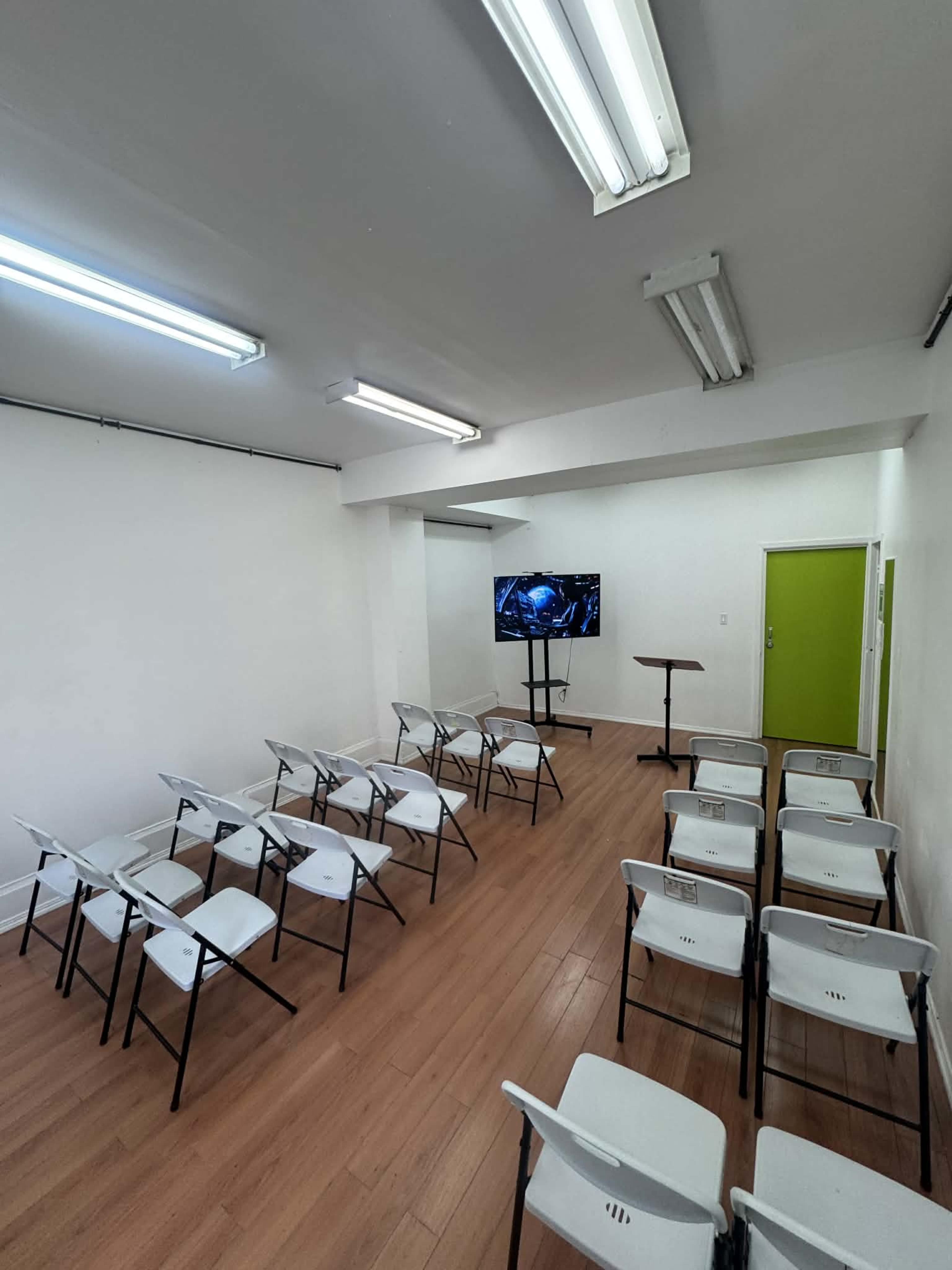 The image shows a classroom setup with several white folding chairs arranged in rows facing a screen on a stand.
