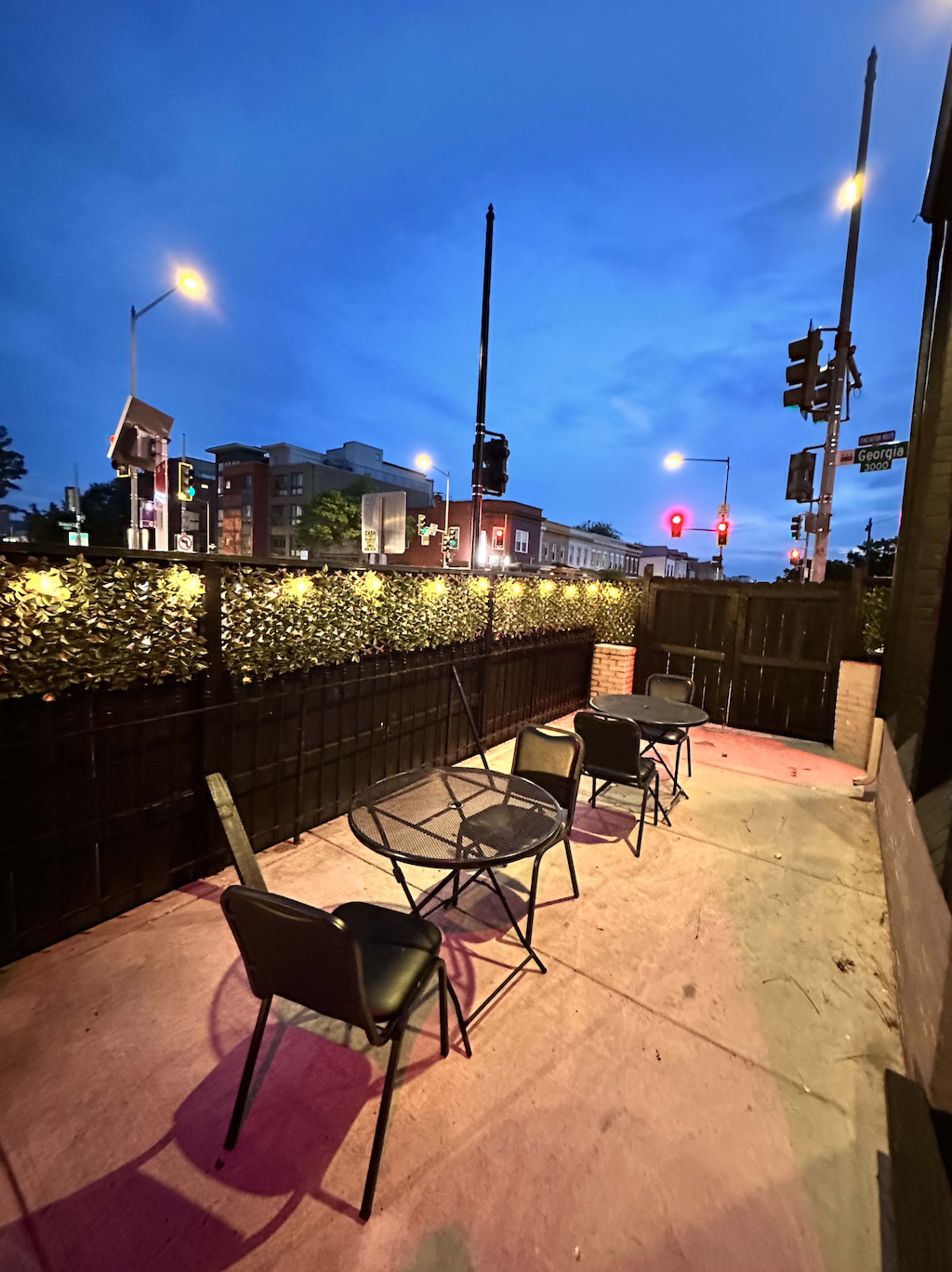 The image shows an empty outdoor seating area at dusk, featuring a few black chairs and a table along a sidewalk.