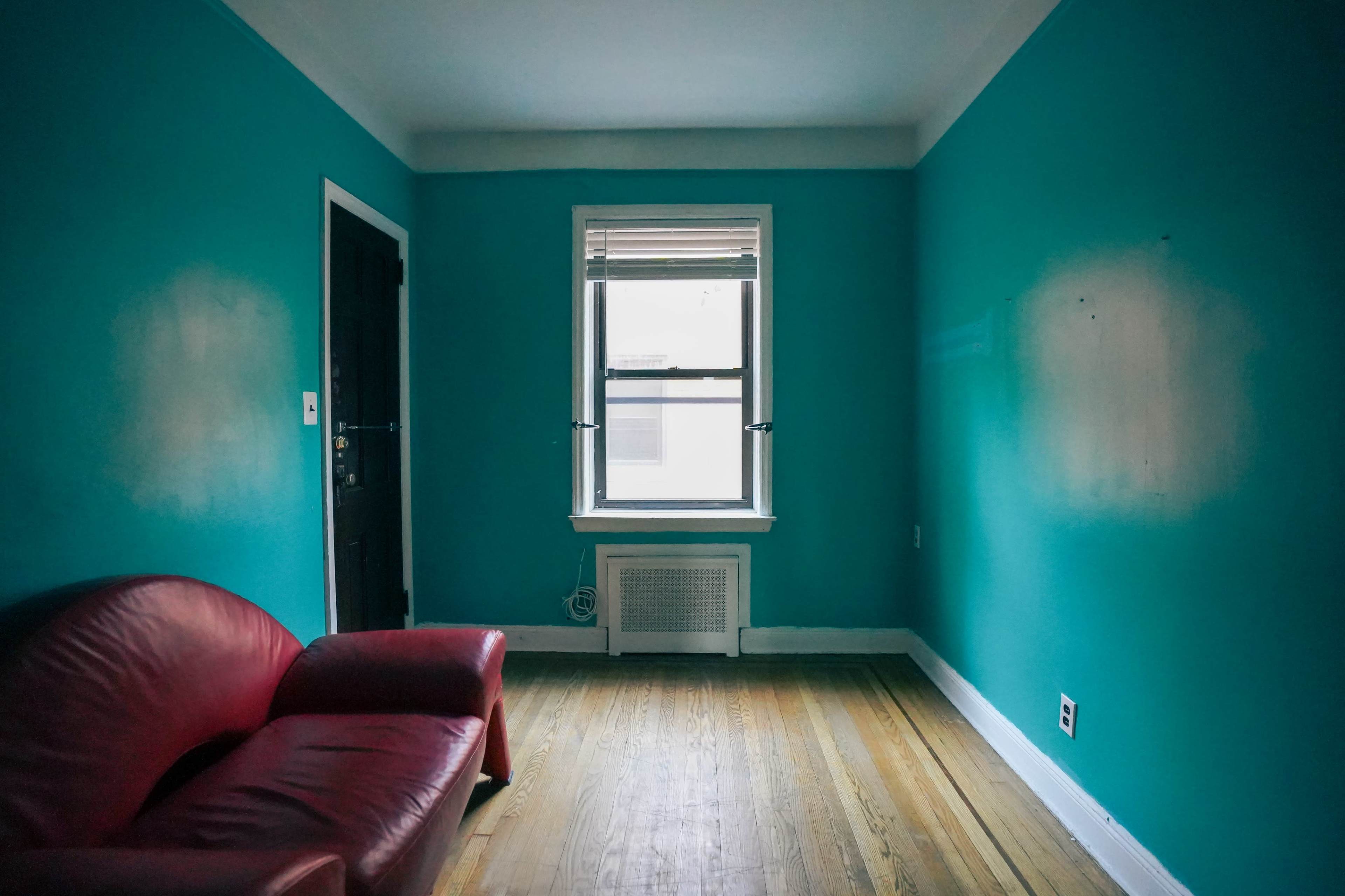 The image shows a sparsely furnished room with teal walls, a red sofa, and a single window.