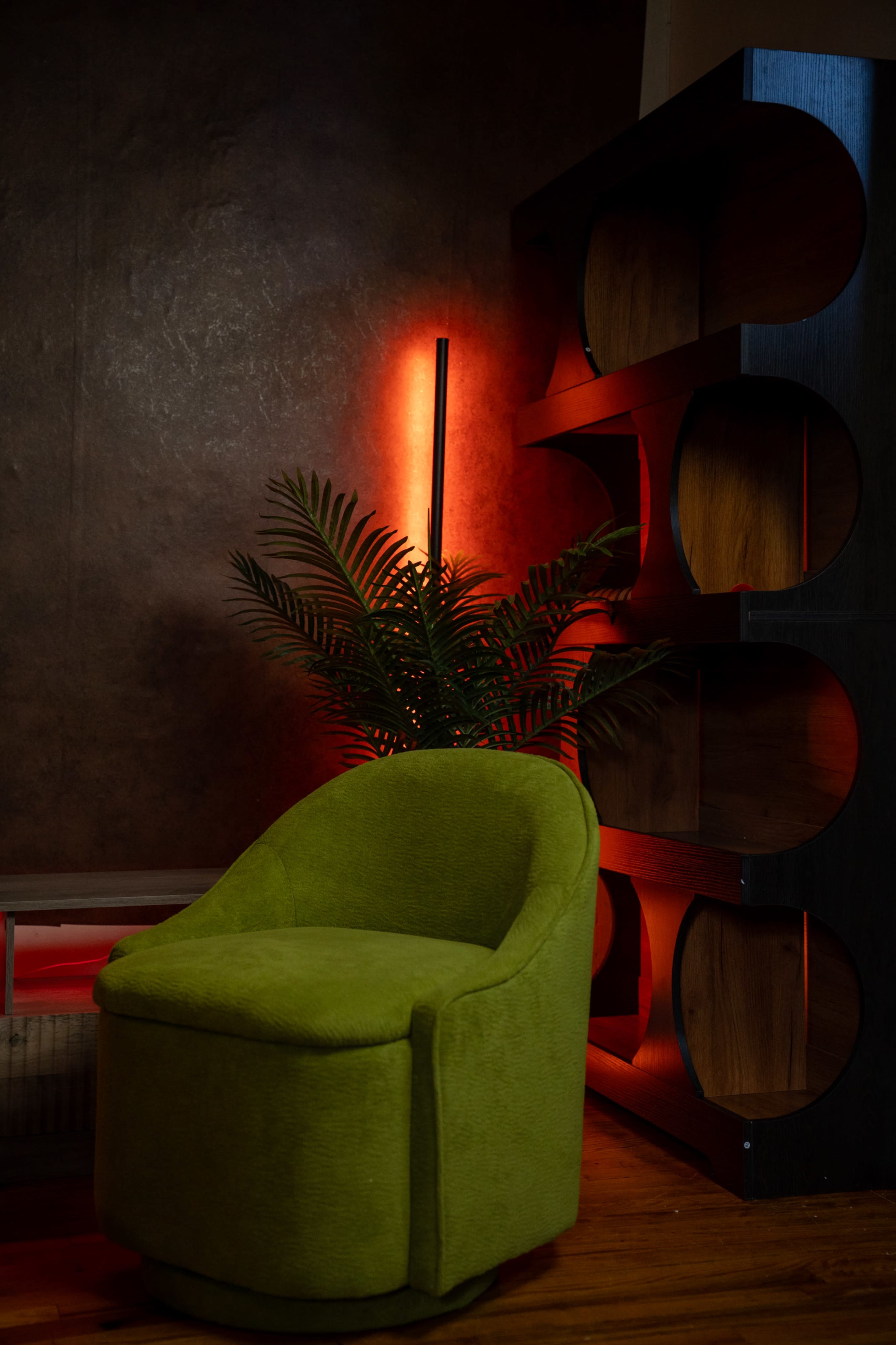 A green swivel chair is positioned in front of a bookshelf with a dark finish, while a tropical plant and red lighting create a striking backdrop.
