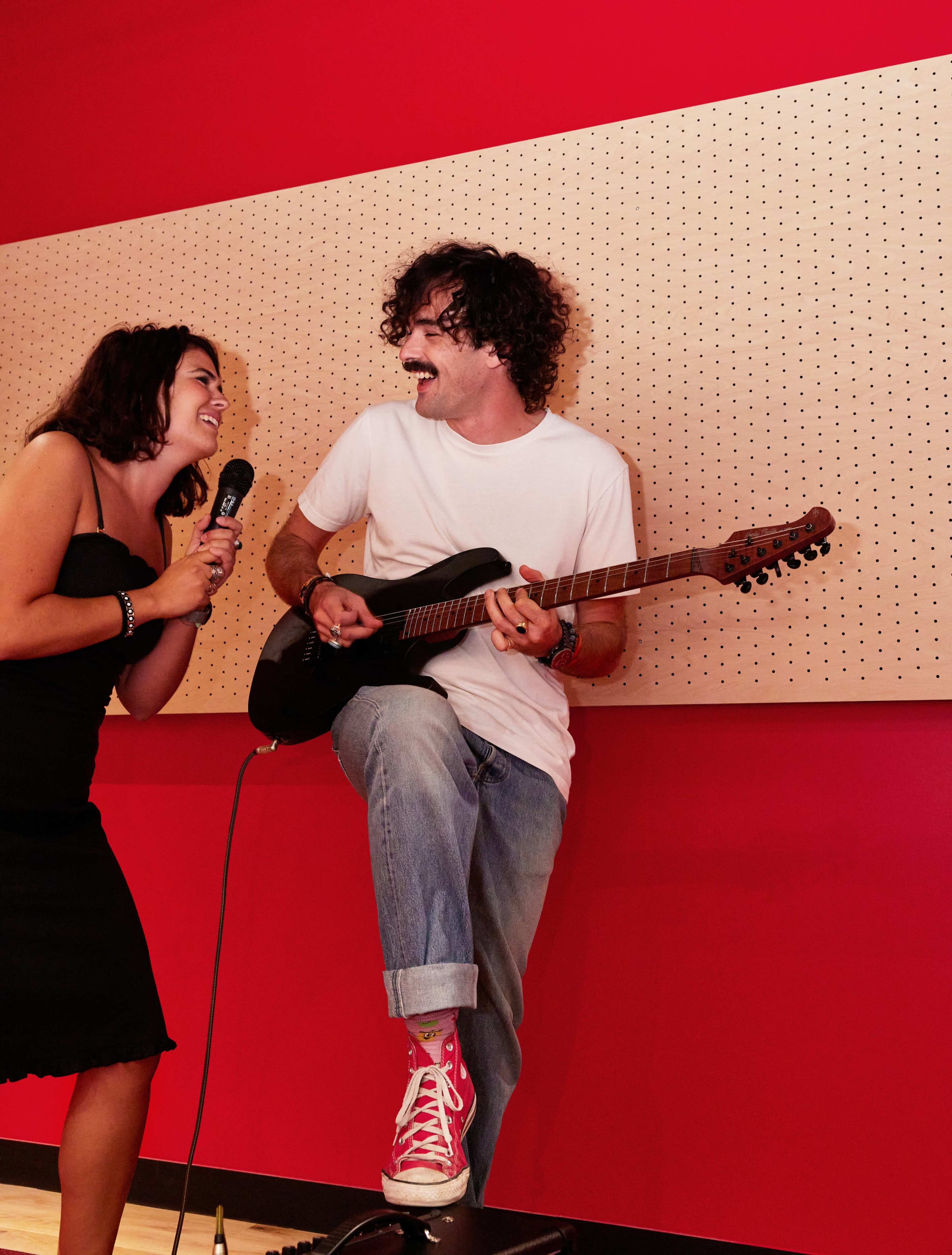 A woman holds a microphone while singing to a man seated with a guitar in a room with red walls.
