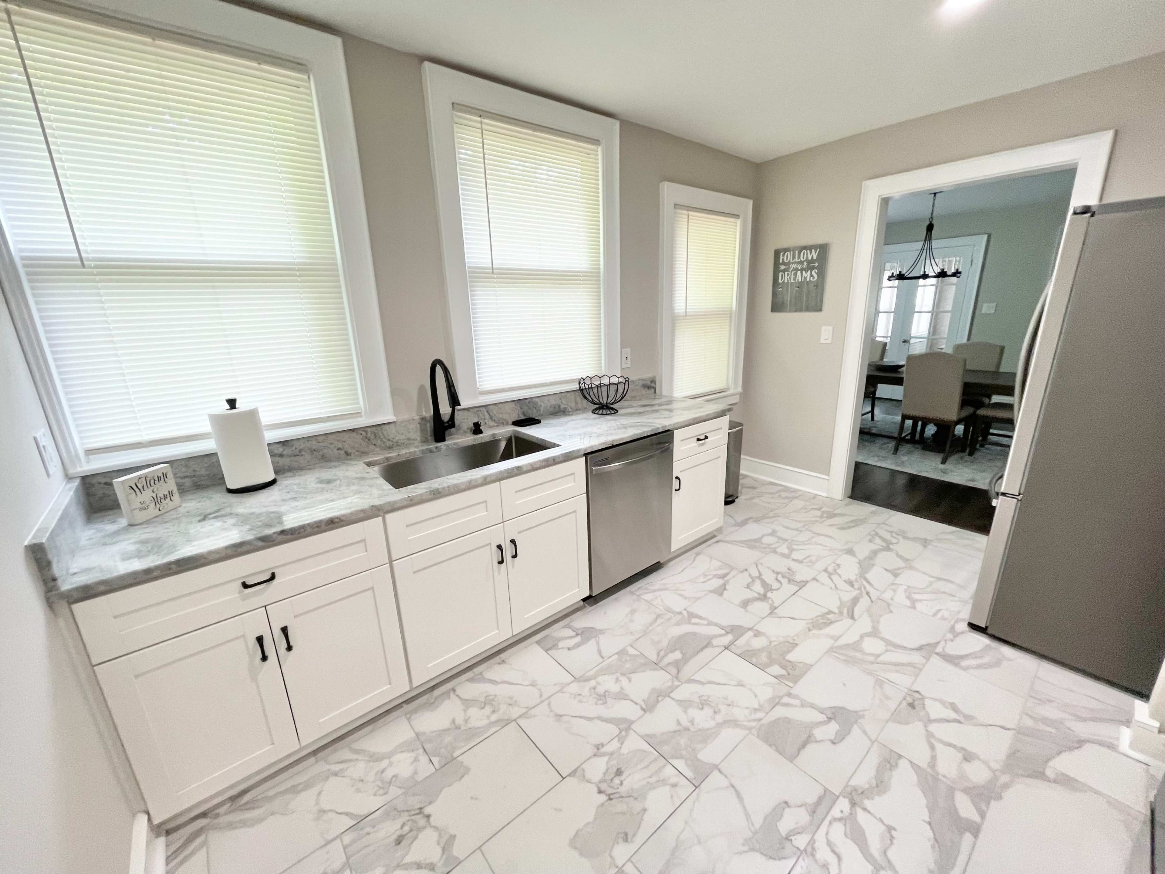 A kitchen features white cabinetry, a stainless steel dishwasher, and a countertop with a marble design, illuminated by natural light from windows.