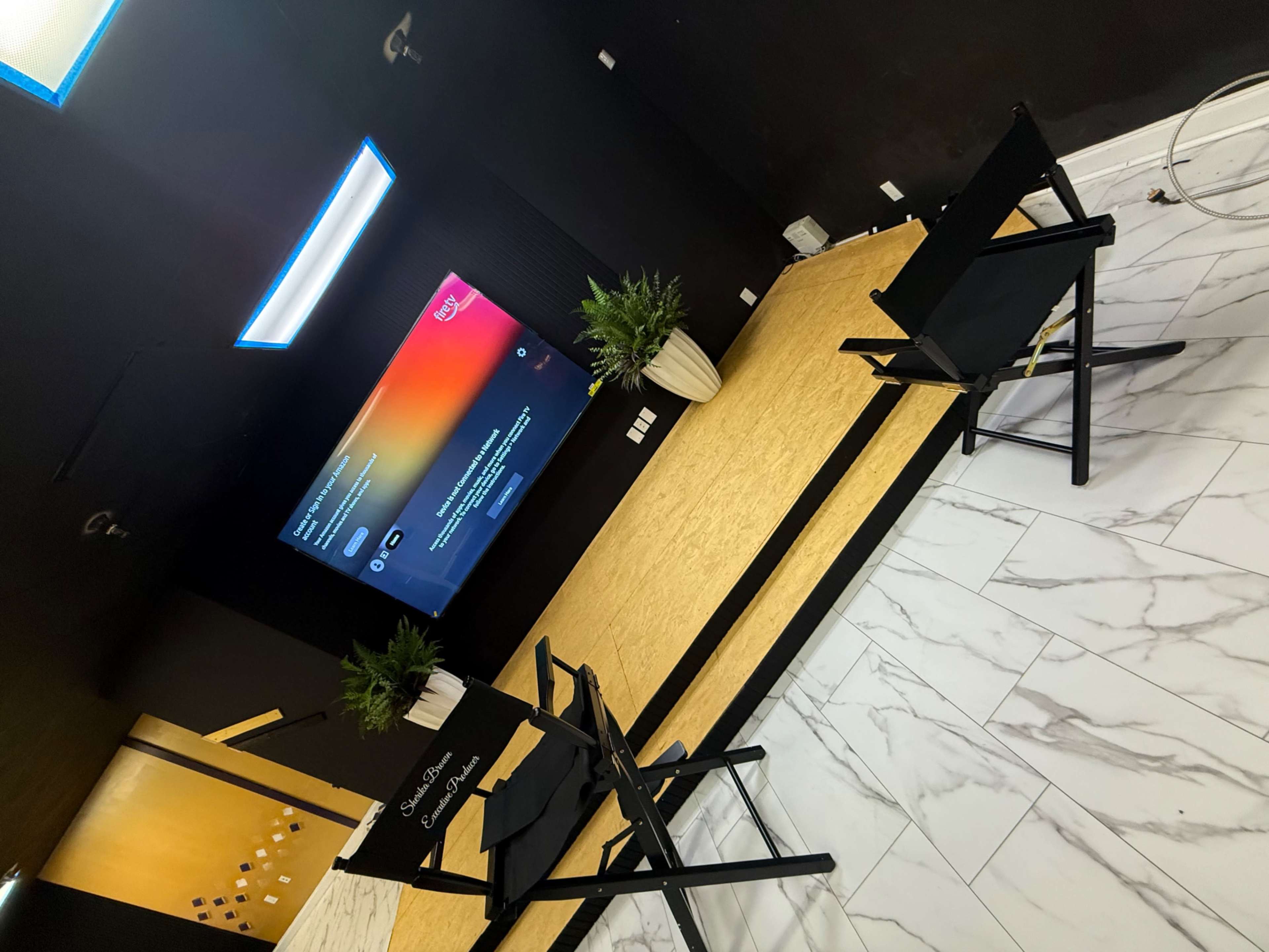 The image shows a modern room with black walls, a large screen displaying a colorful interface, and two black director's chairs facing the screen, with small potted plants on either side.