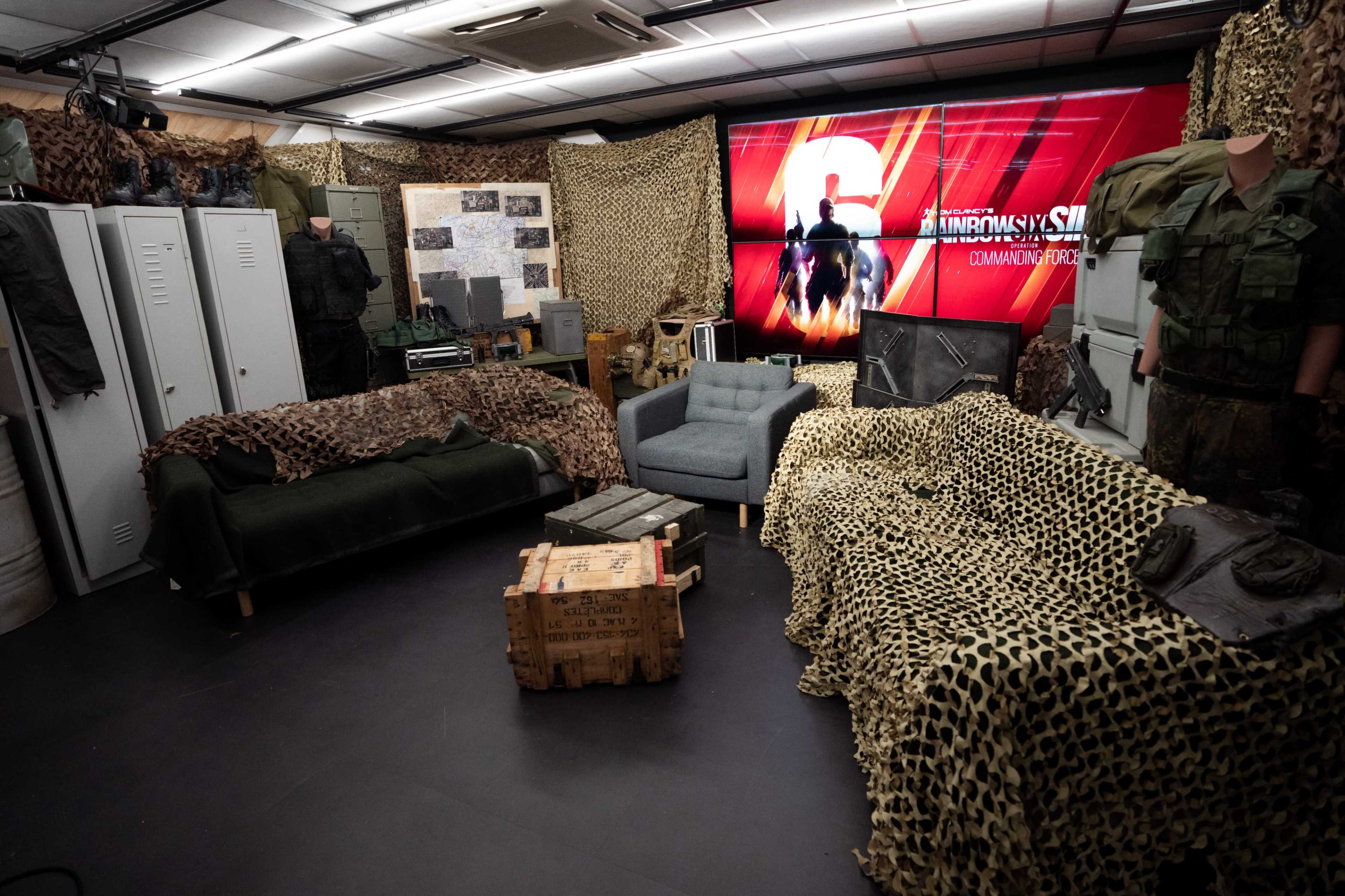 The scene depicts a room decorated with military camouflage netting, featuring seating, military uniforms displayed on mannequins, and a large screen showing a graphic.