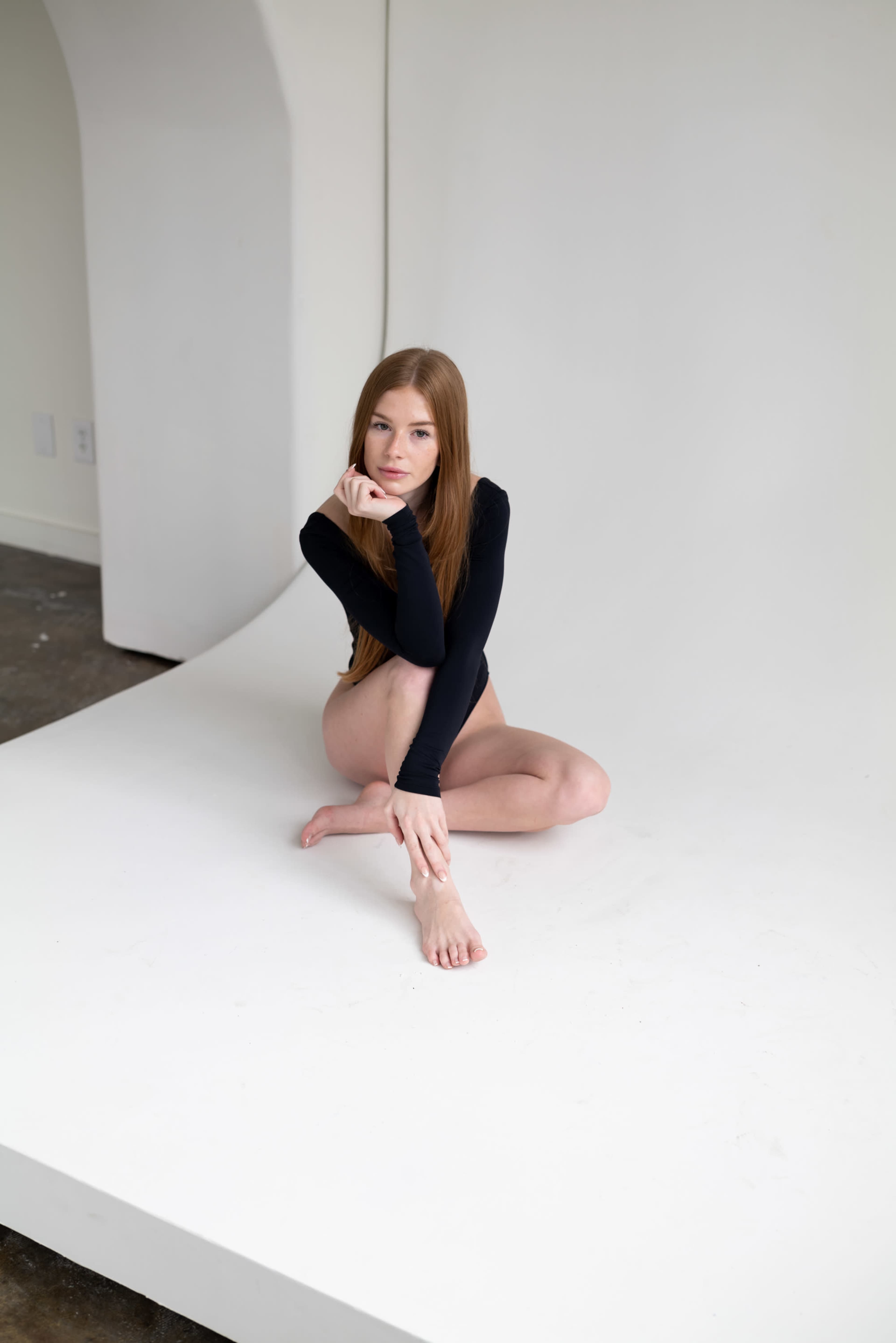 A person with long hair sits on a seamless white backdrop, positioned with one arm resting on their knee.