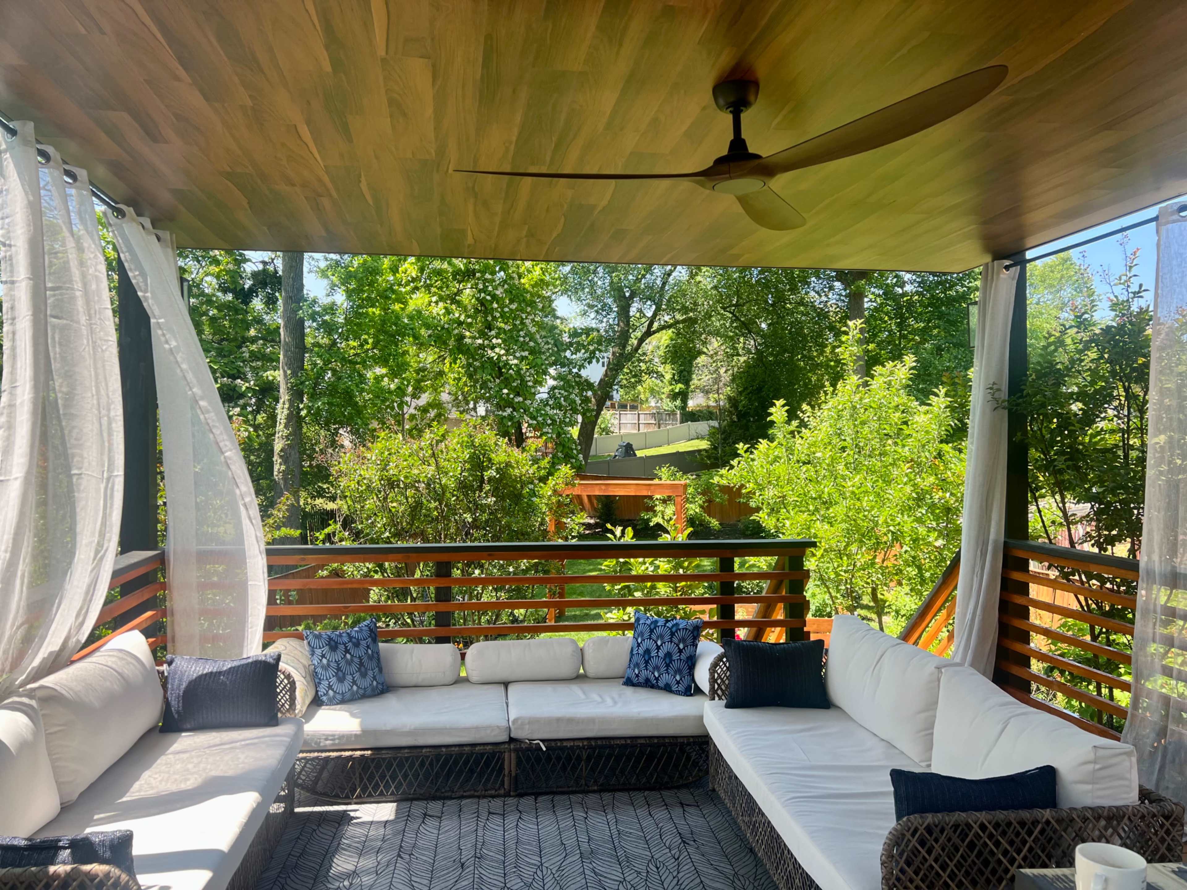 A covered outdoor seating area with a sectional sofa, decorative pillows, and a fan, surrounded by greenery.