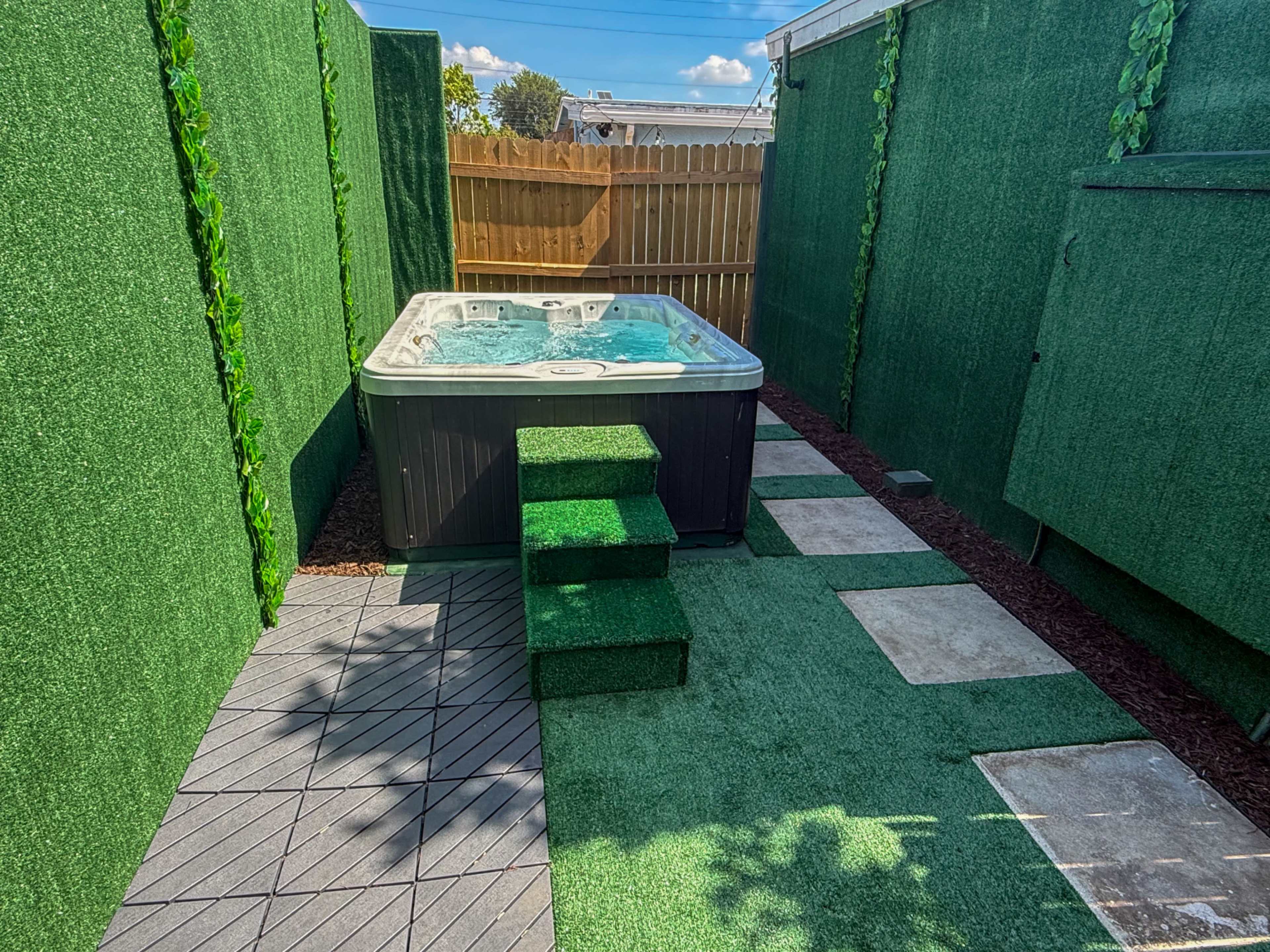 The image shows a backyard area with a hot tub surrounded by artificial grass and wooden fencing.