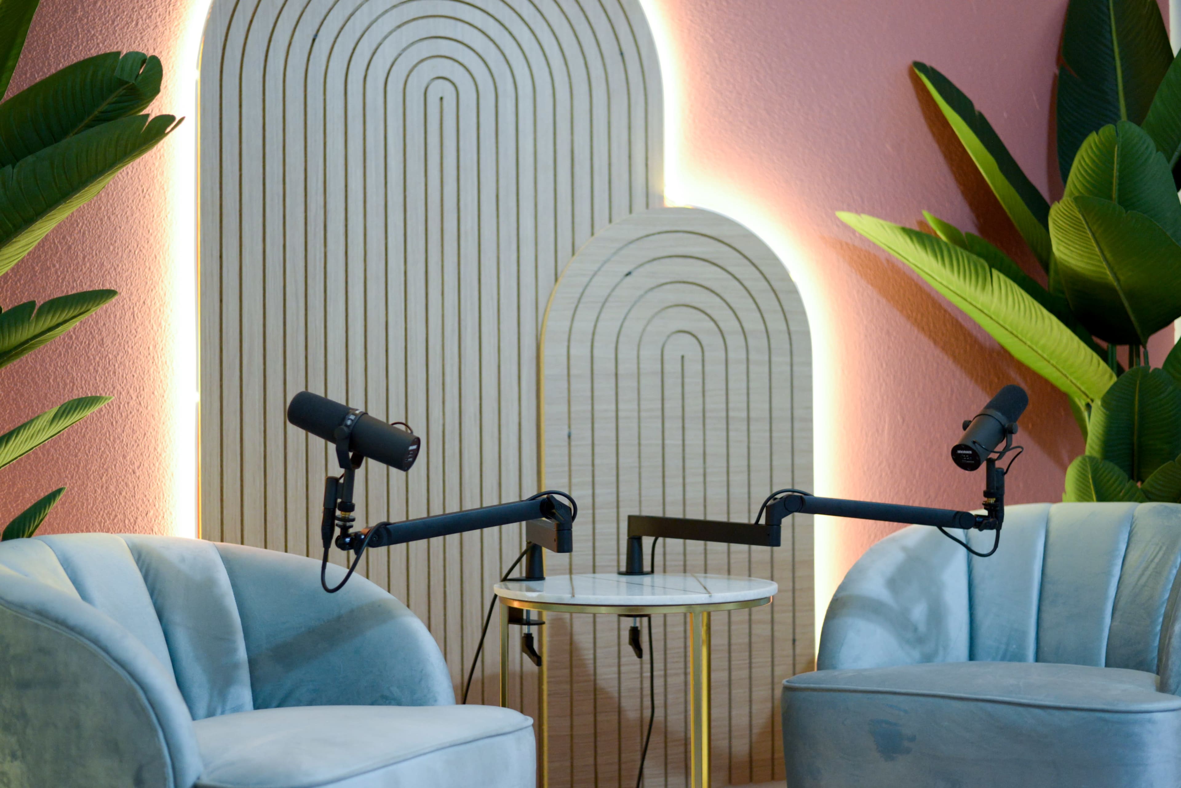 The image shows two microphones positioned on adjustable arms facing two blue velvet chairs beside a round table, set against a backdrop of wooden arch designs and green plants.