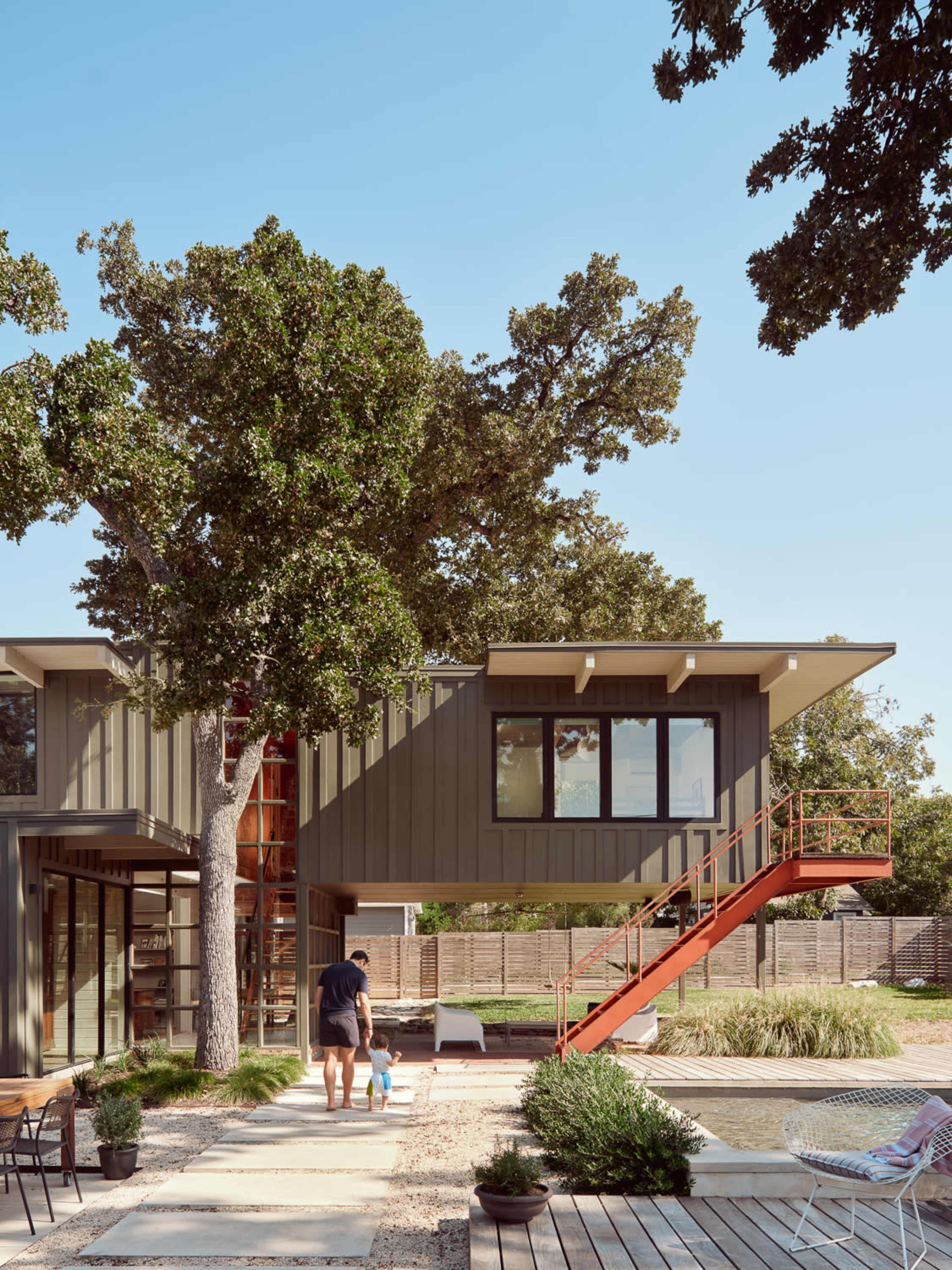 A person and a child walk towards a modern two-story house with a metal exterior and an external staircase, surrounded by greenery and landscaped paths.