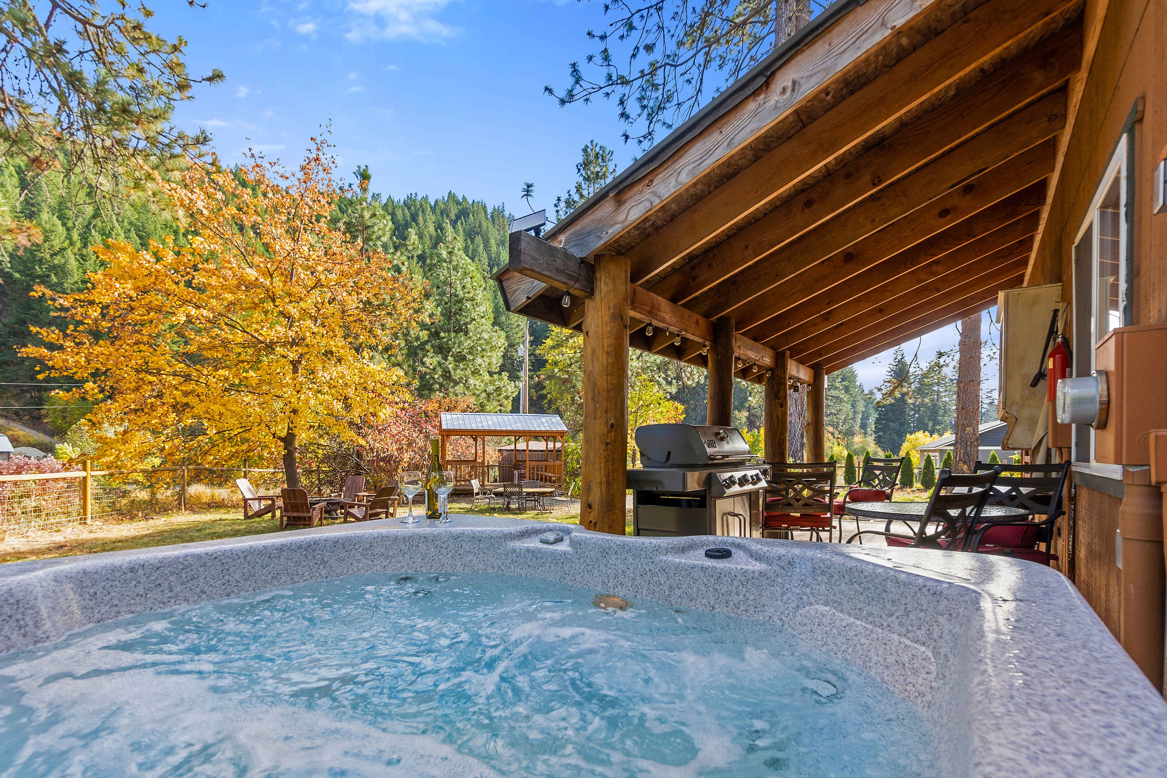 The image shows a hot tub on a wooden deck, surrounded by trees with autumn foliage and a barbecue grill nearby.