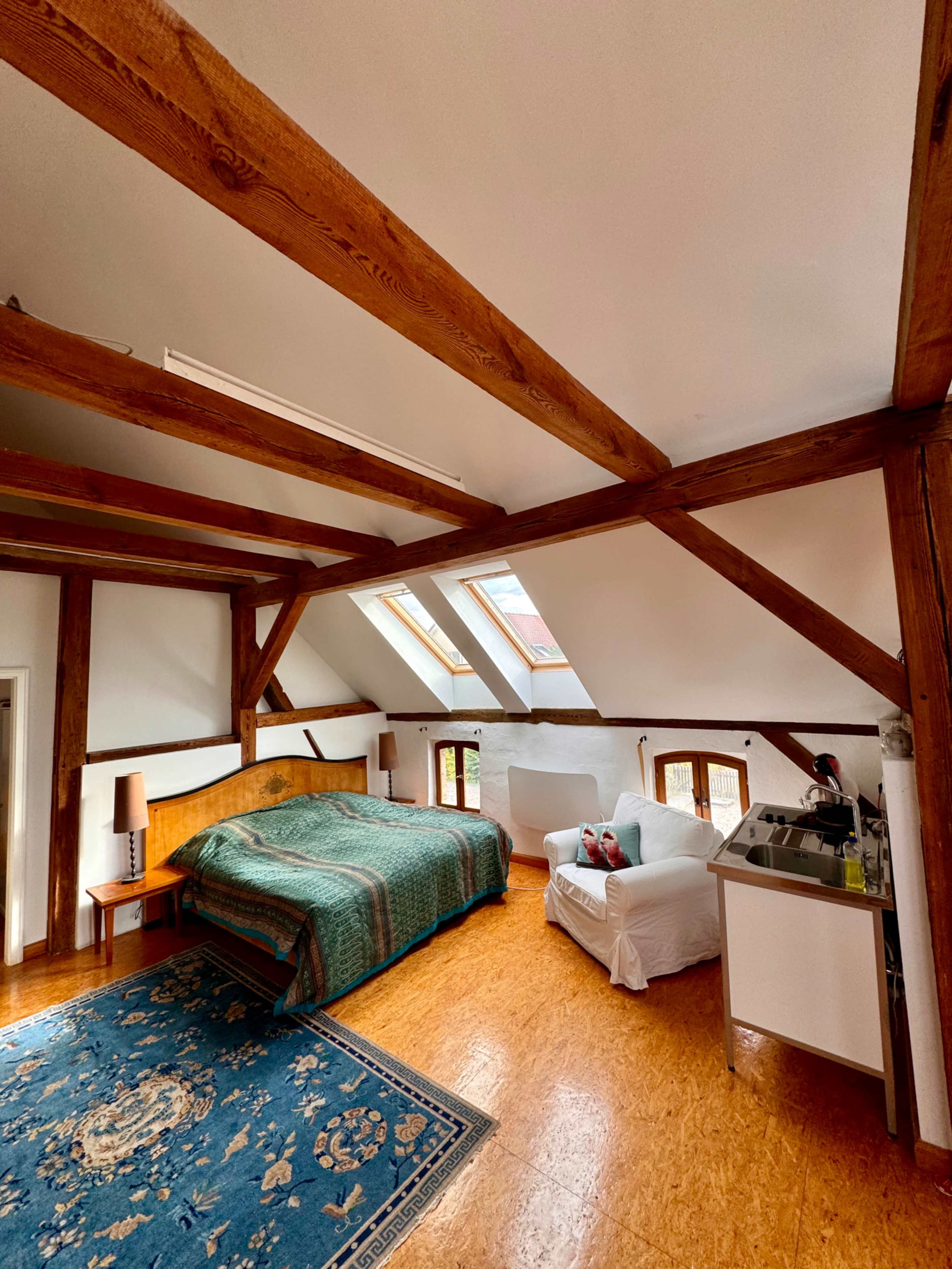 The image shows a spacious bedroom with wooden beams, a large bed, a blue area rug, a white armchair, and a small kitchen area in one corner.