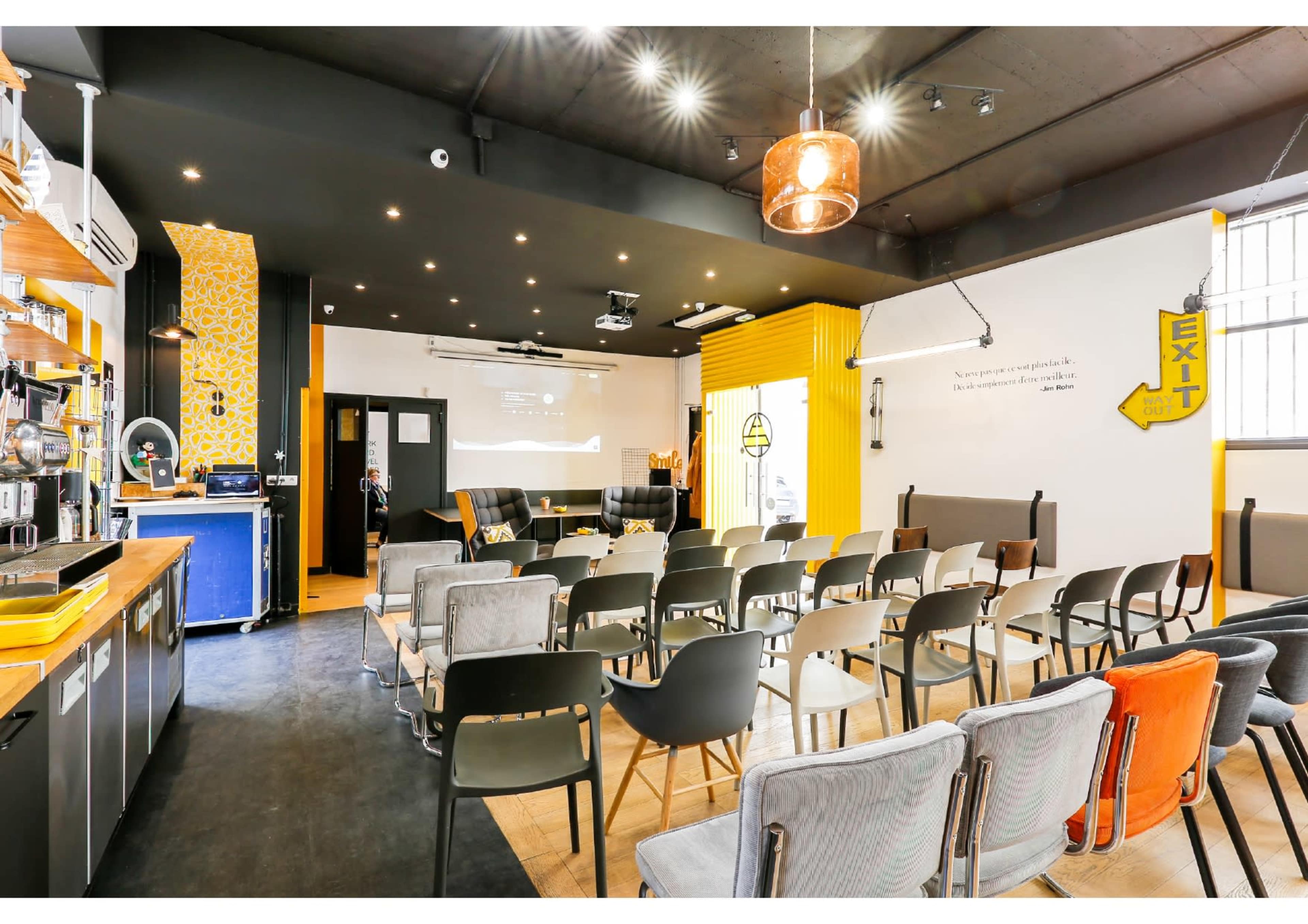 A modern workshop space features rows of chairs facing a presentation area, with a coffee bar and bright yellow accents on the walls.