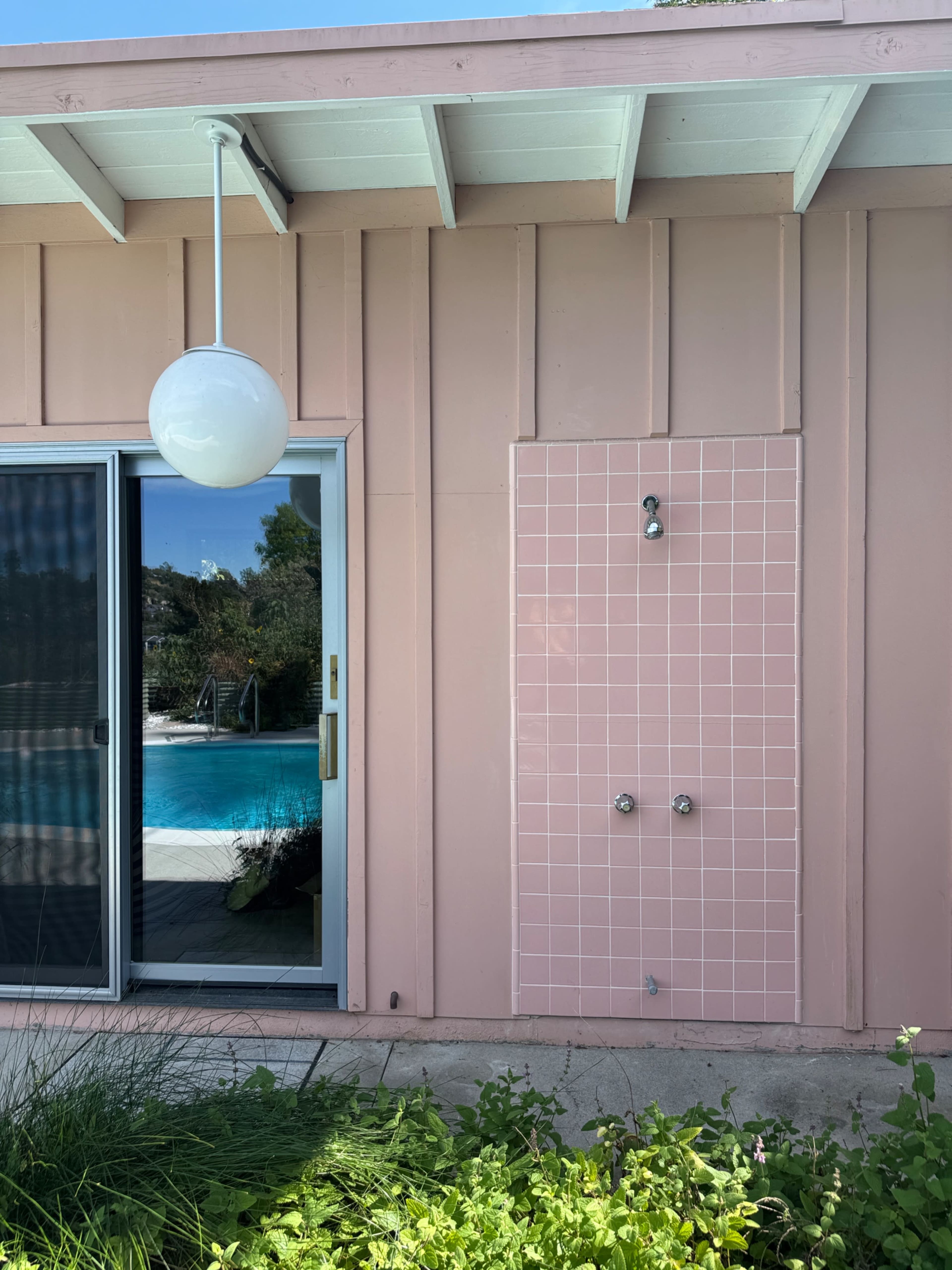 The image shows a pink tiled outdoor shower next to a glass sliding door that leads to a swimming pool area.