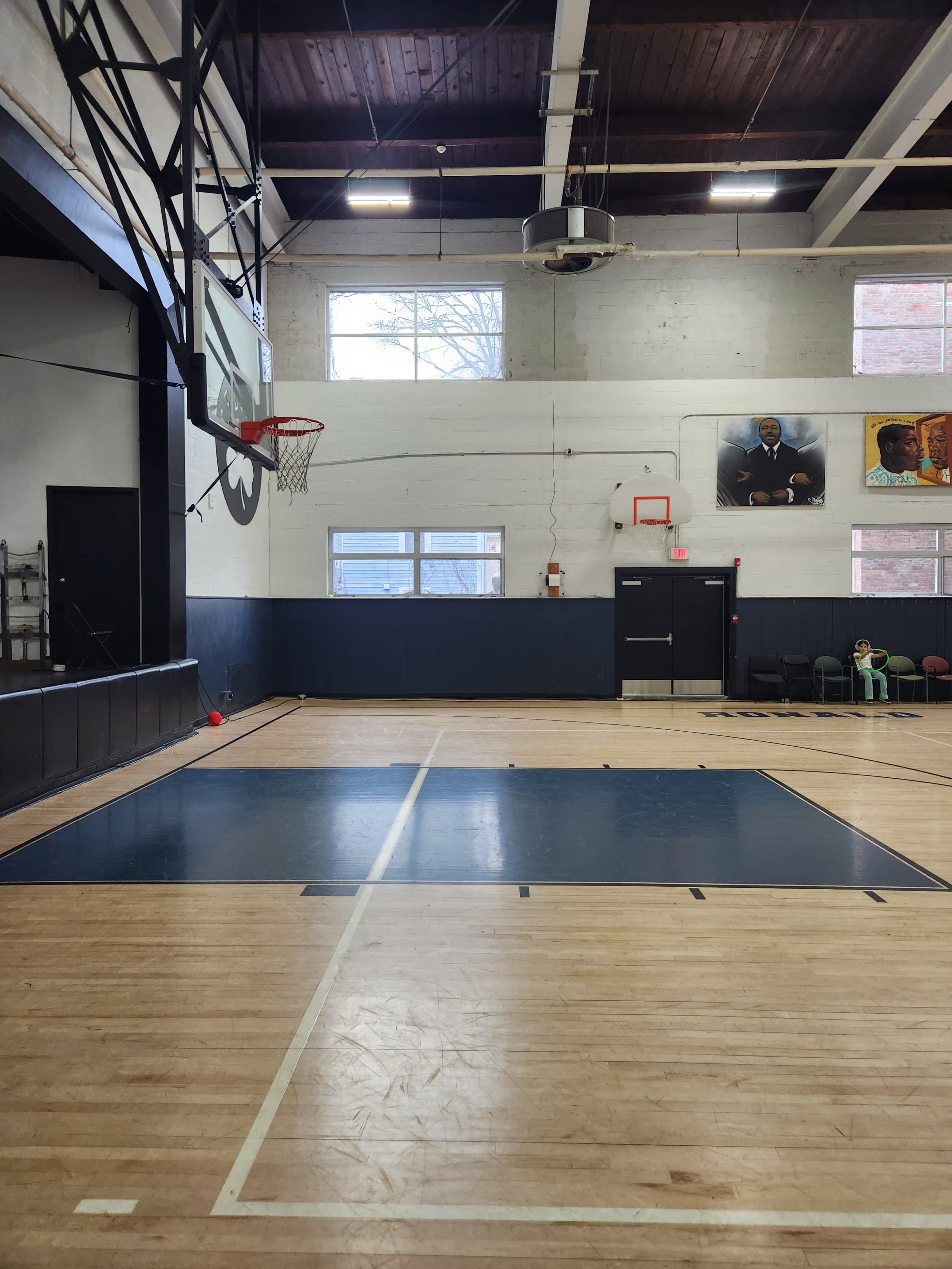 The image shows a basketball court featuring a hoop, hardwood flooring, and large windows alongside wall murals.