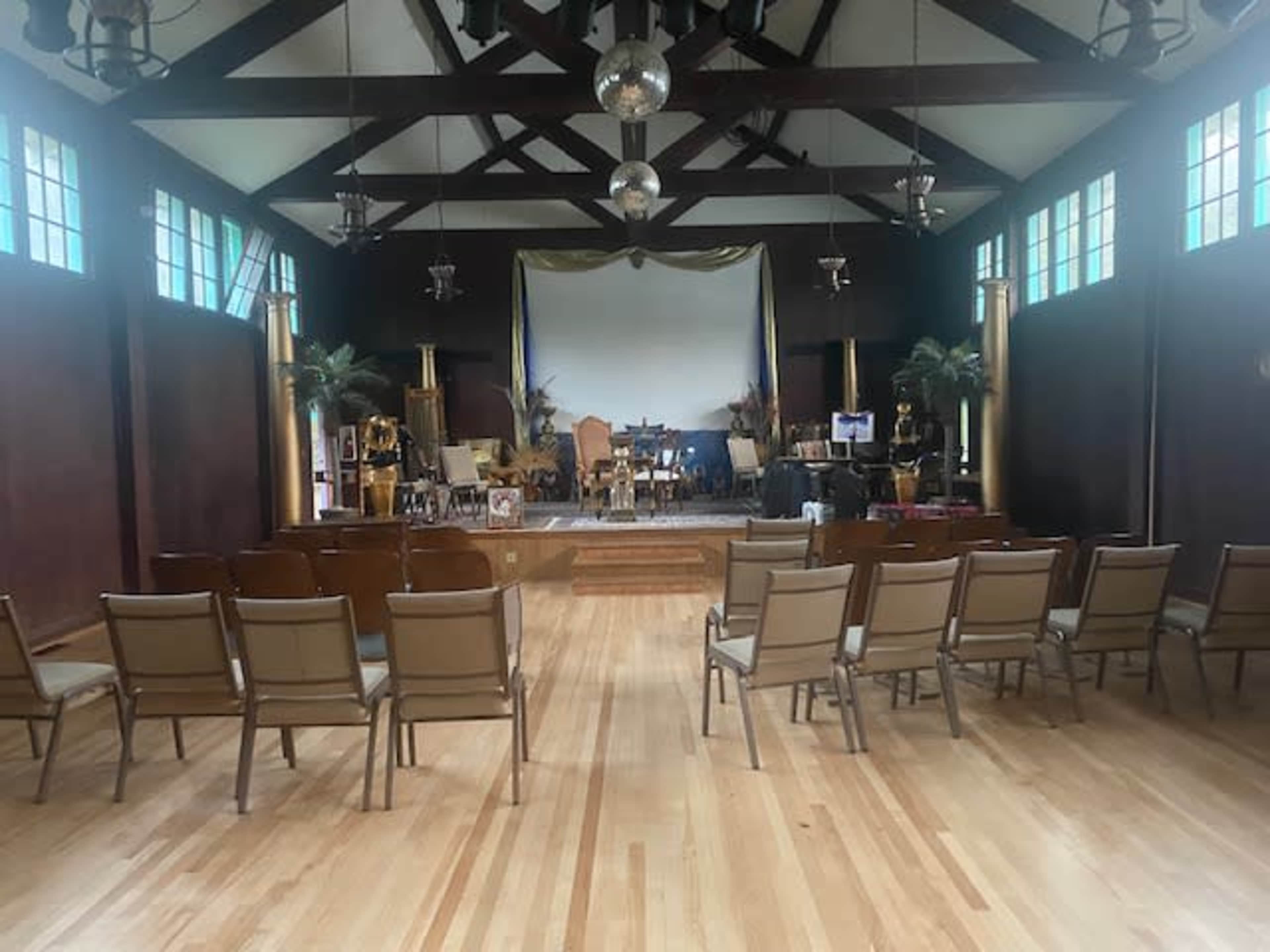 A spacious indoor hall with wooden flooring features rows of chairs facing a stage adorned with musical instruments and decorative plants.