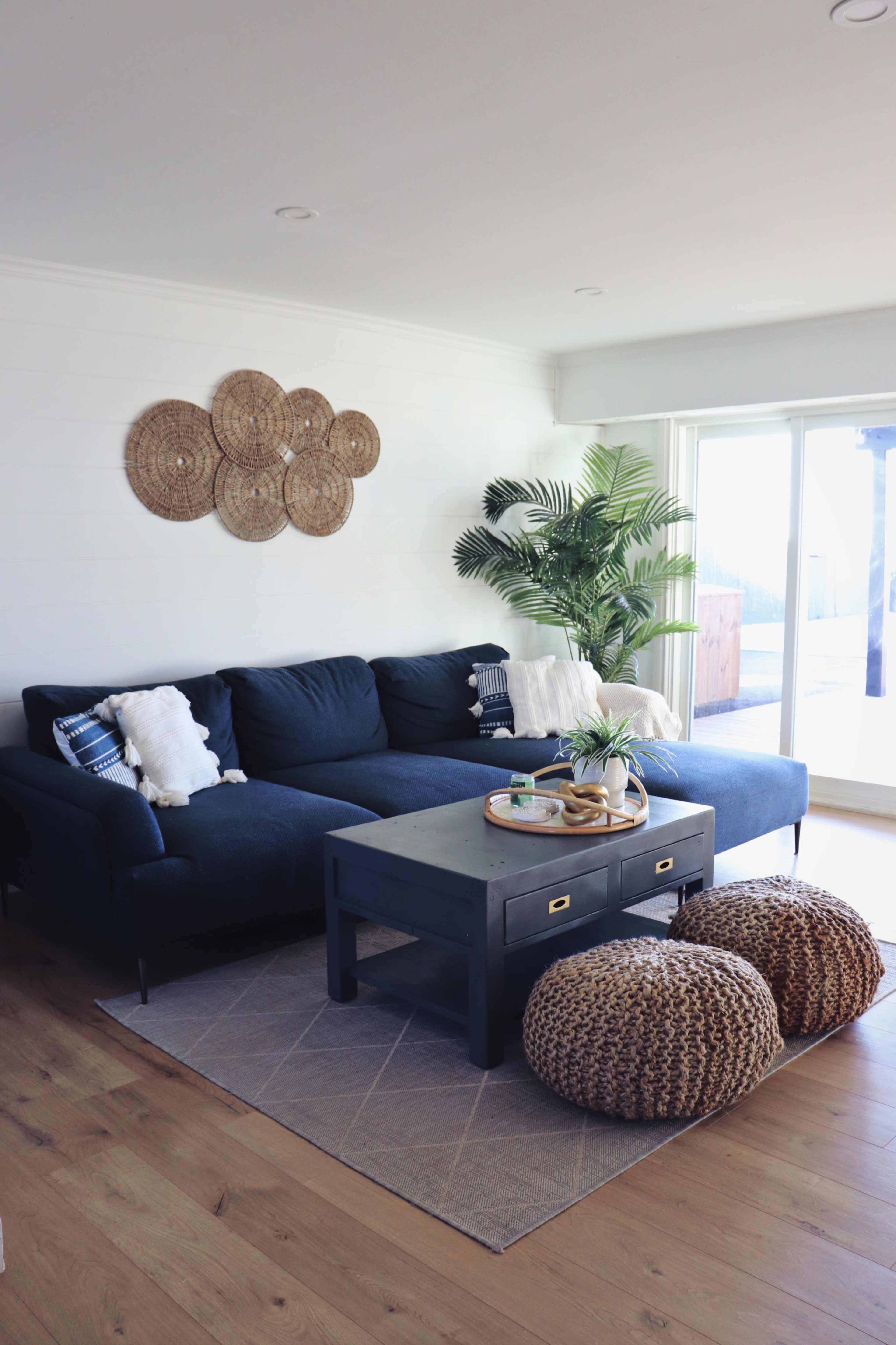 A modern living room features a dark blue sectional sofa, a wooden coffee table, and woven poufs, along with decorative wall art and a potted plant.