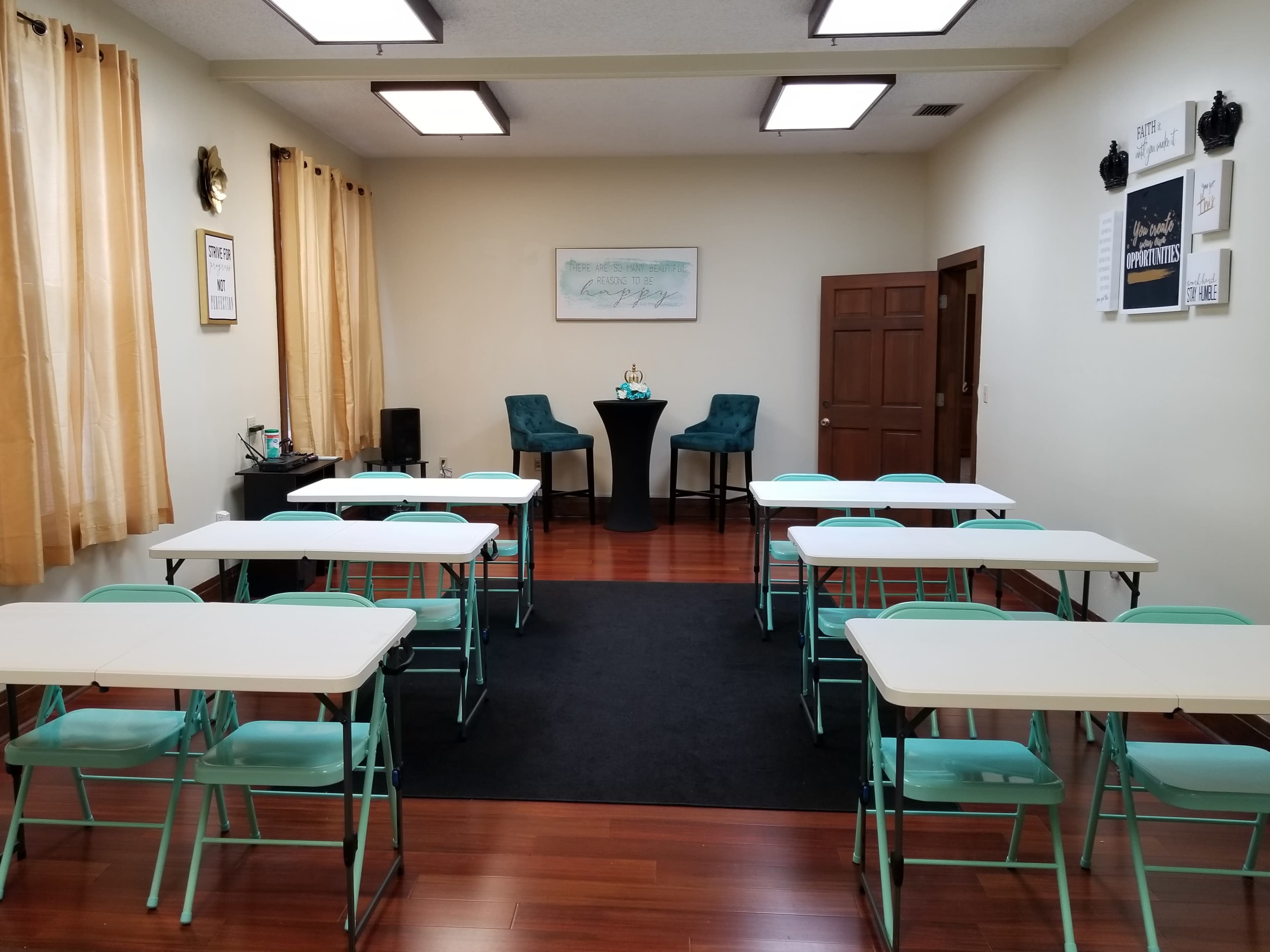 The room features several rows of white tables and teal chairs, with a small table set up at the front and large windows adorned with yellow curtains.