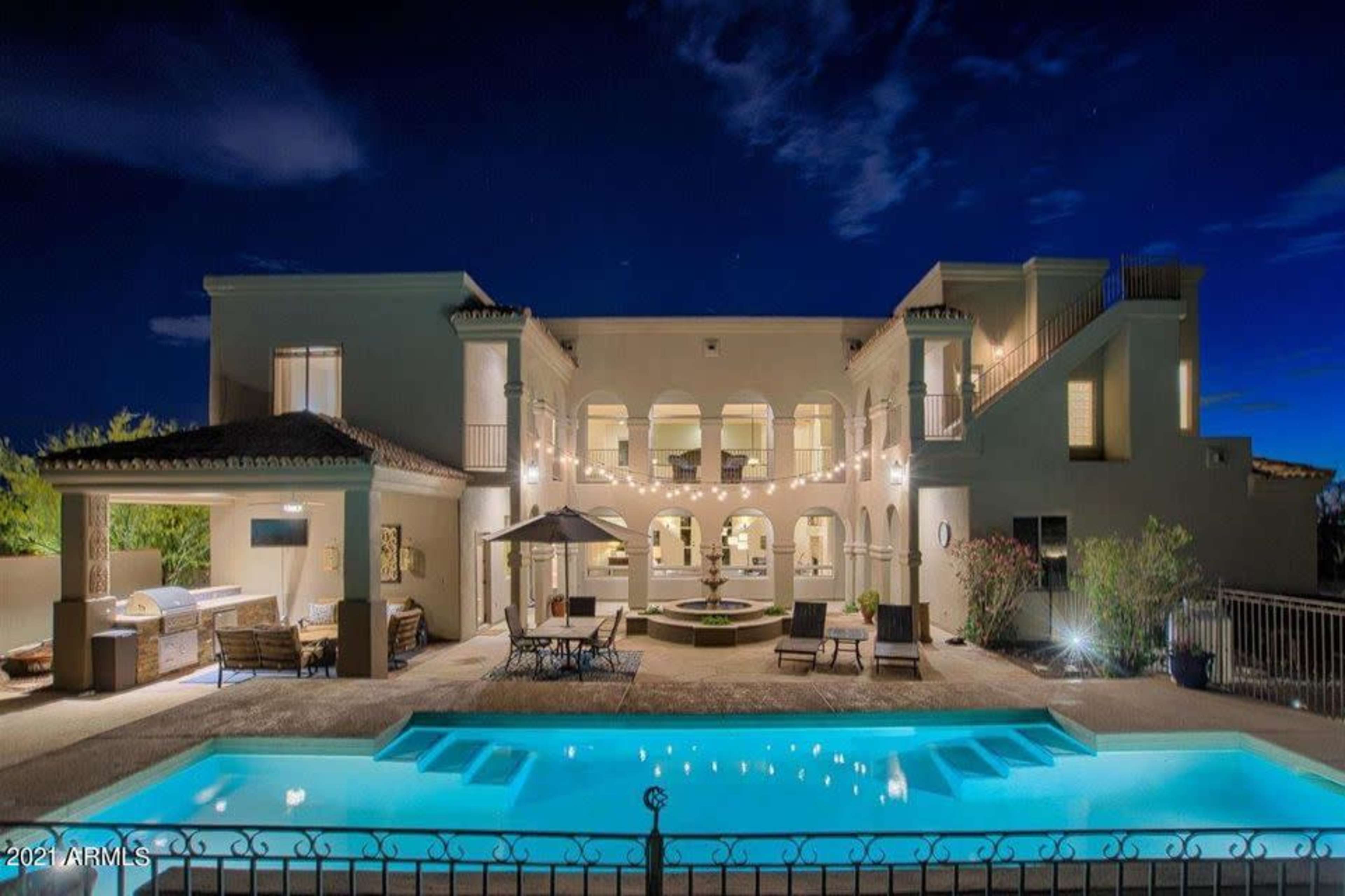 A large, illuminated house with multiple arches, a swimming pool in the foreground, and outdoor seating areas under a clear night sky.