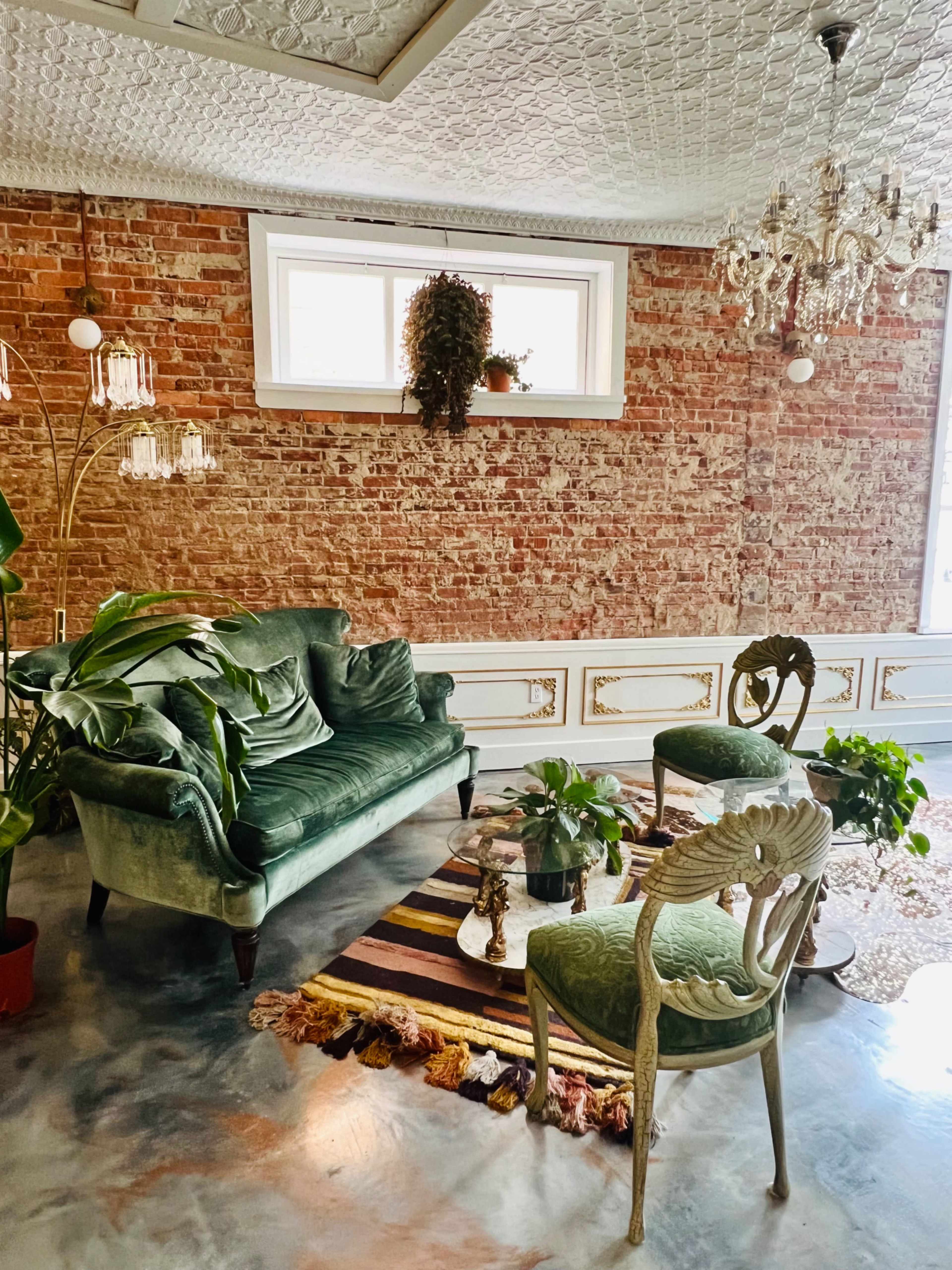A living room with exposed brick walls, a green velvet sofa, a decorative armchair, and a patterned rug, all surrounded by various potted plants.