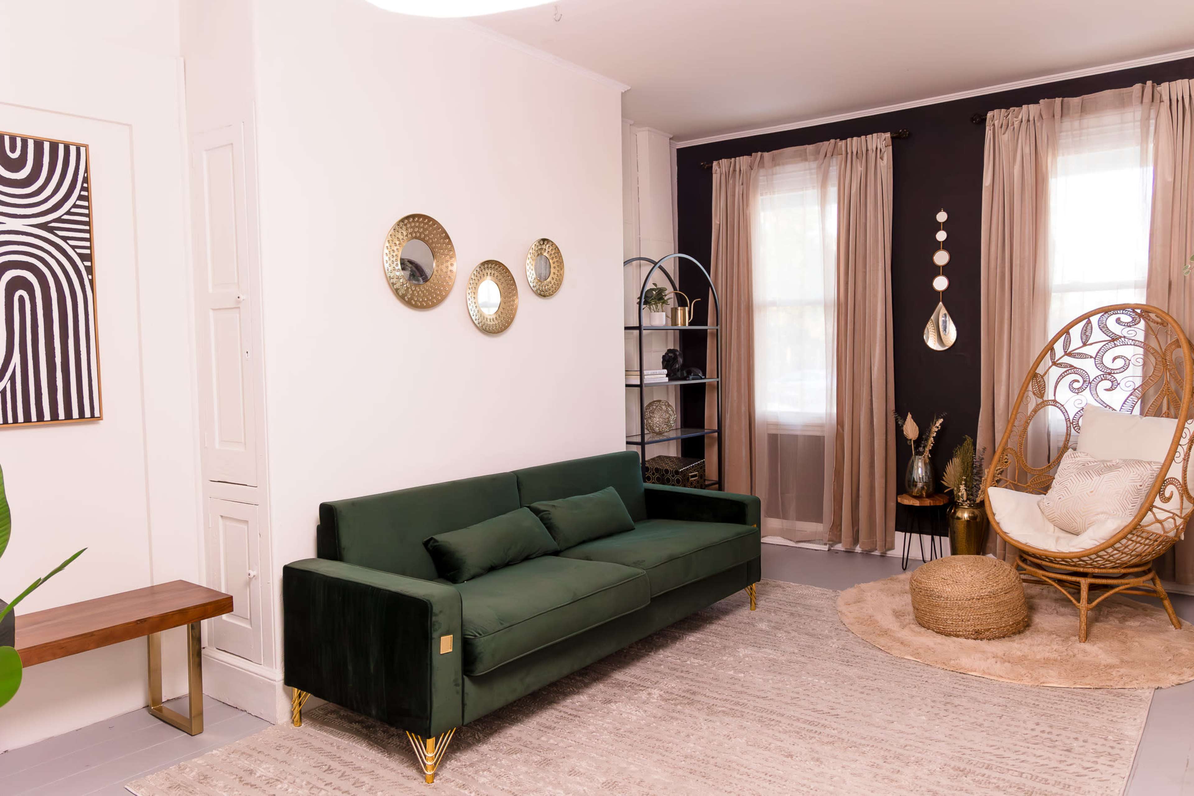 The image shows a living room featuring a green velvet sofa, a round chair made of wicker, decorative mirrors on the wall, and a bookshelf with plants and decor items.