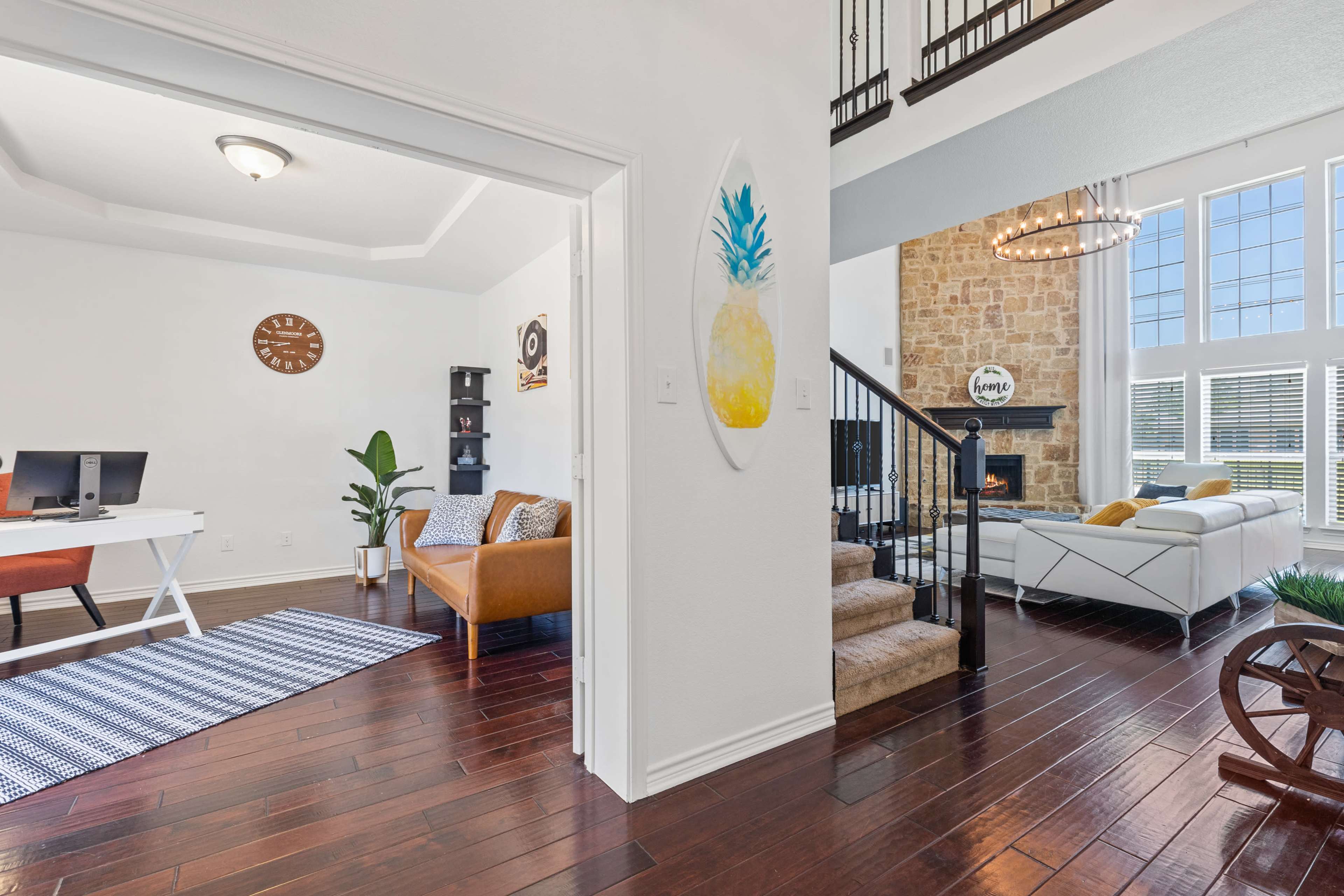 A bright, open living space featuring a home office area with a desk and chair, adjacent to a cozy seating area, and a staircase leading to a second level with large windows and a stone fireplace.