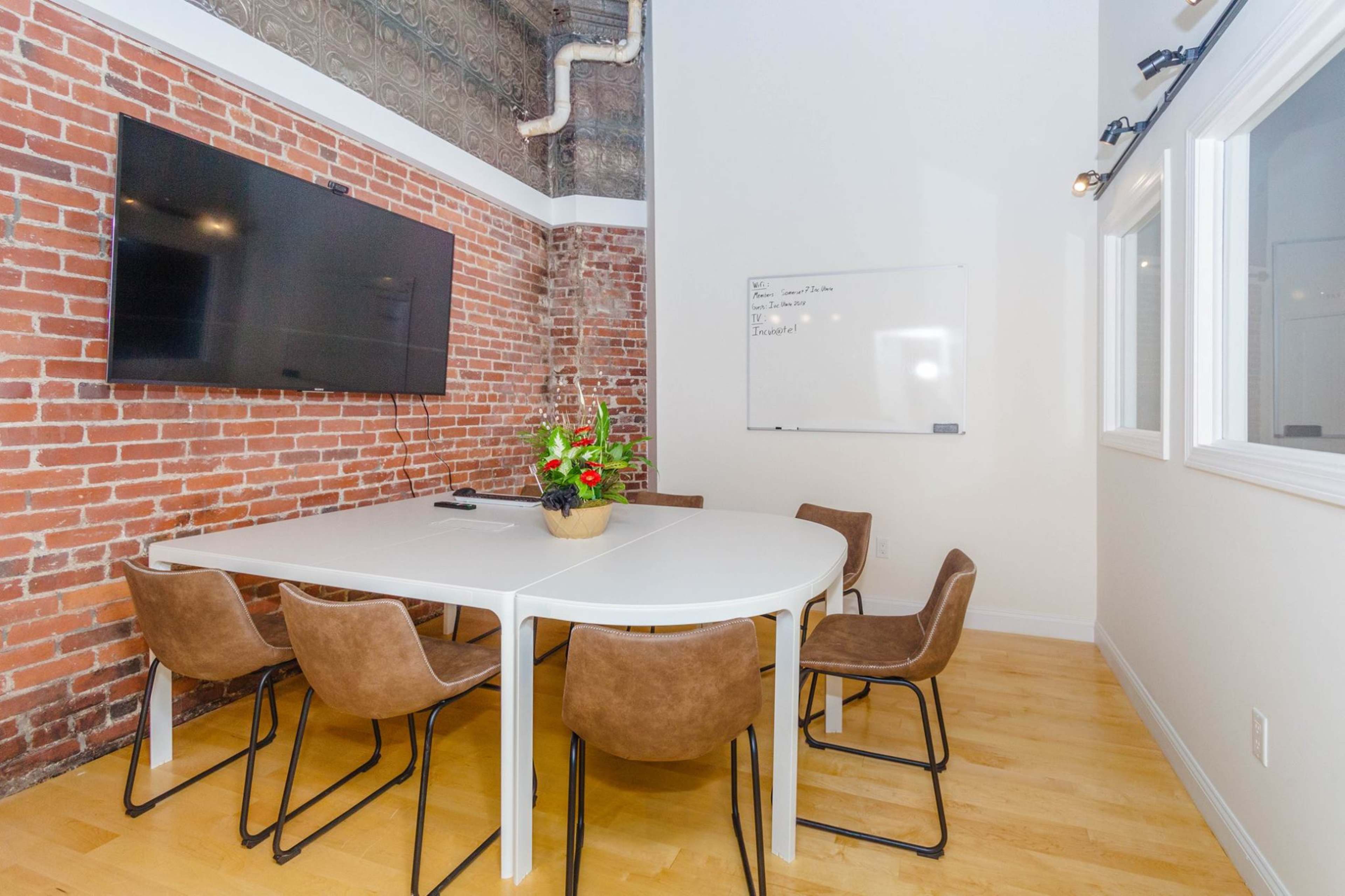 A meeting room features a round table surrounded by chairs, a large wall-mounted television, and a whiteboard on the wall.