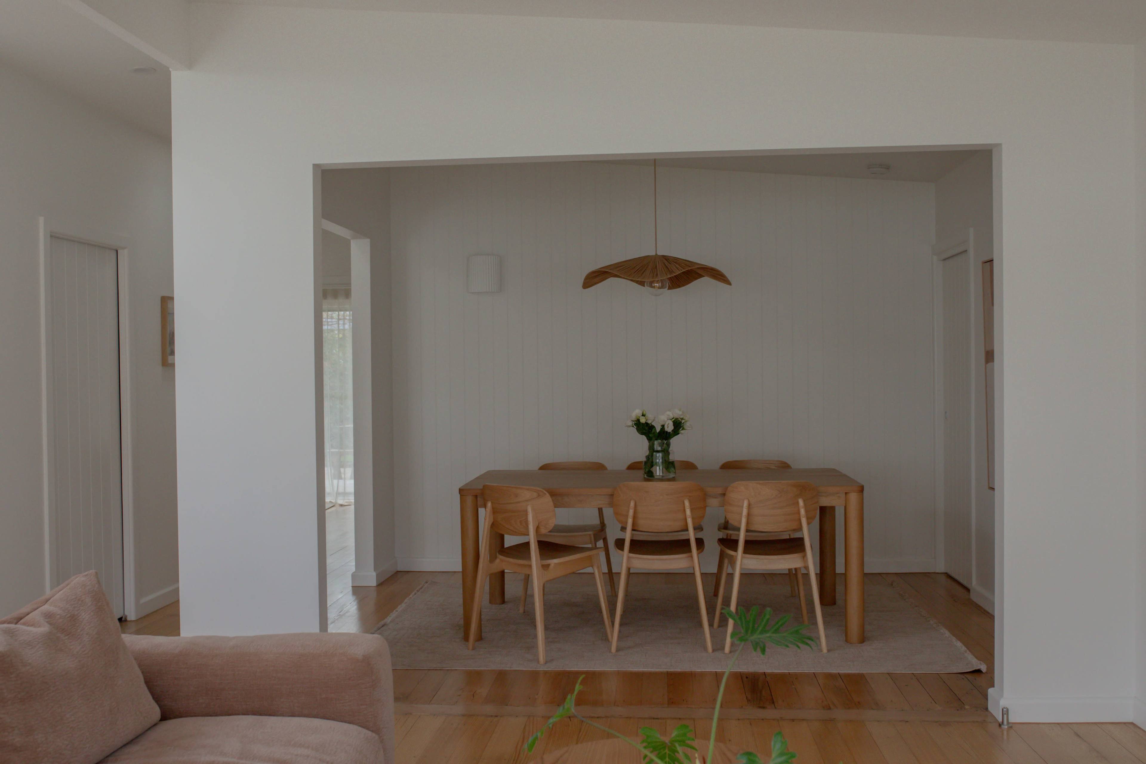 The image shows a modern dining area featuring a wooden table surrounded by chairs, with a vase of flowers as the centerpiece, all set against a light-colored wall.