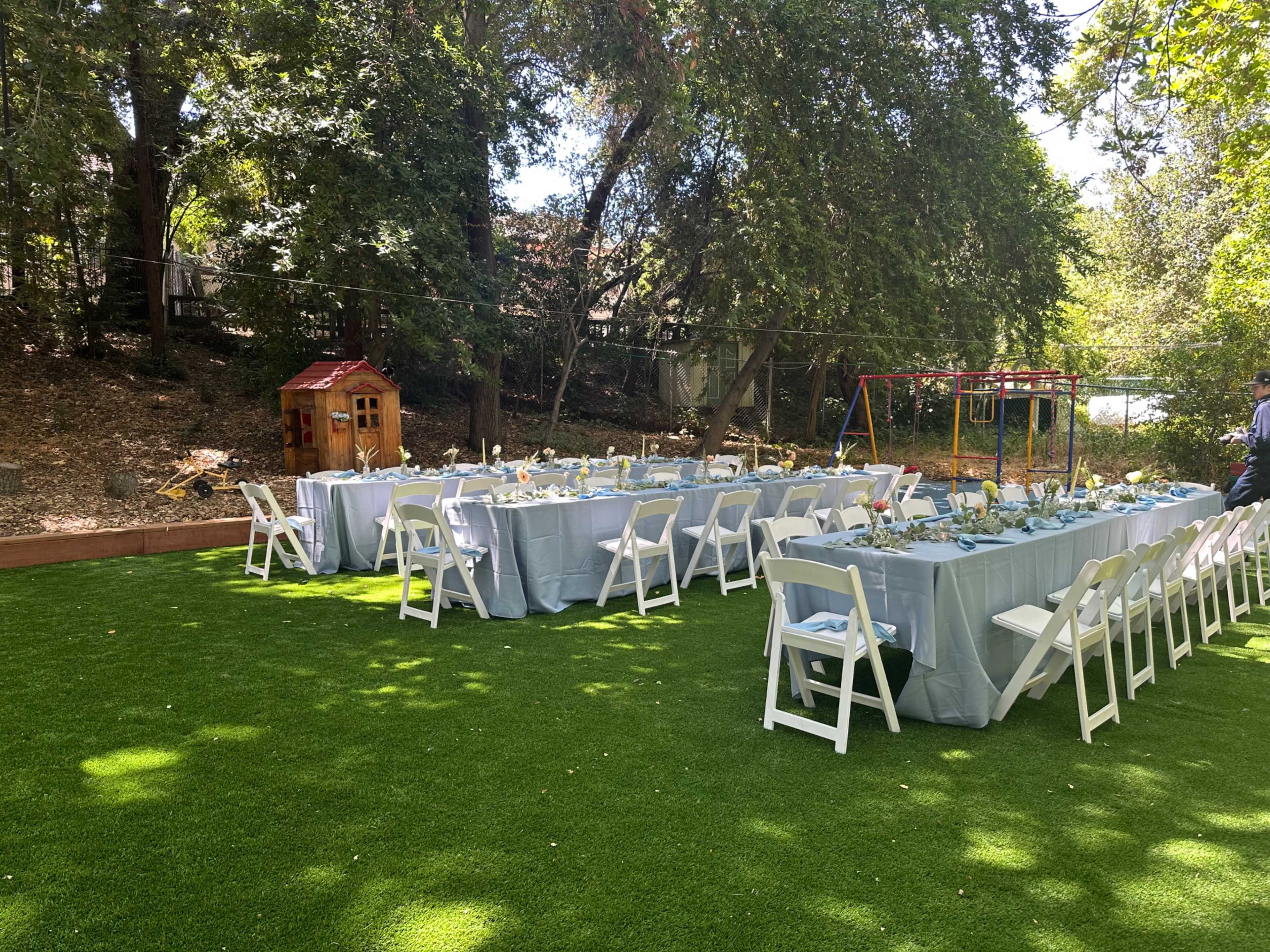 A large outdoor dining setup with long tables and white chairs is arranged on green turf, surrounded by trees and children's play equipment.