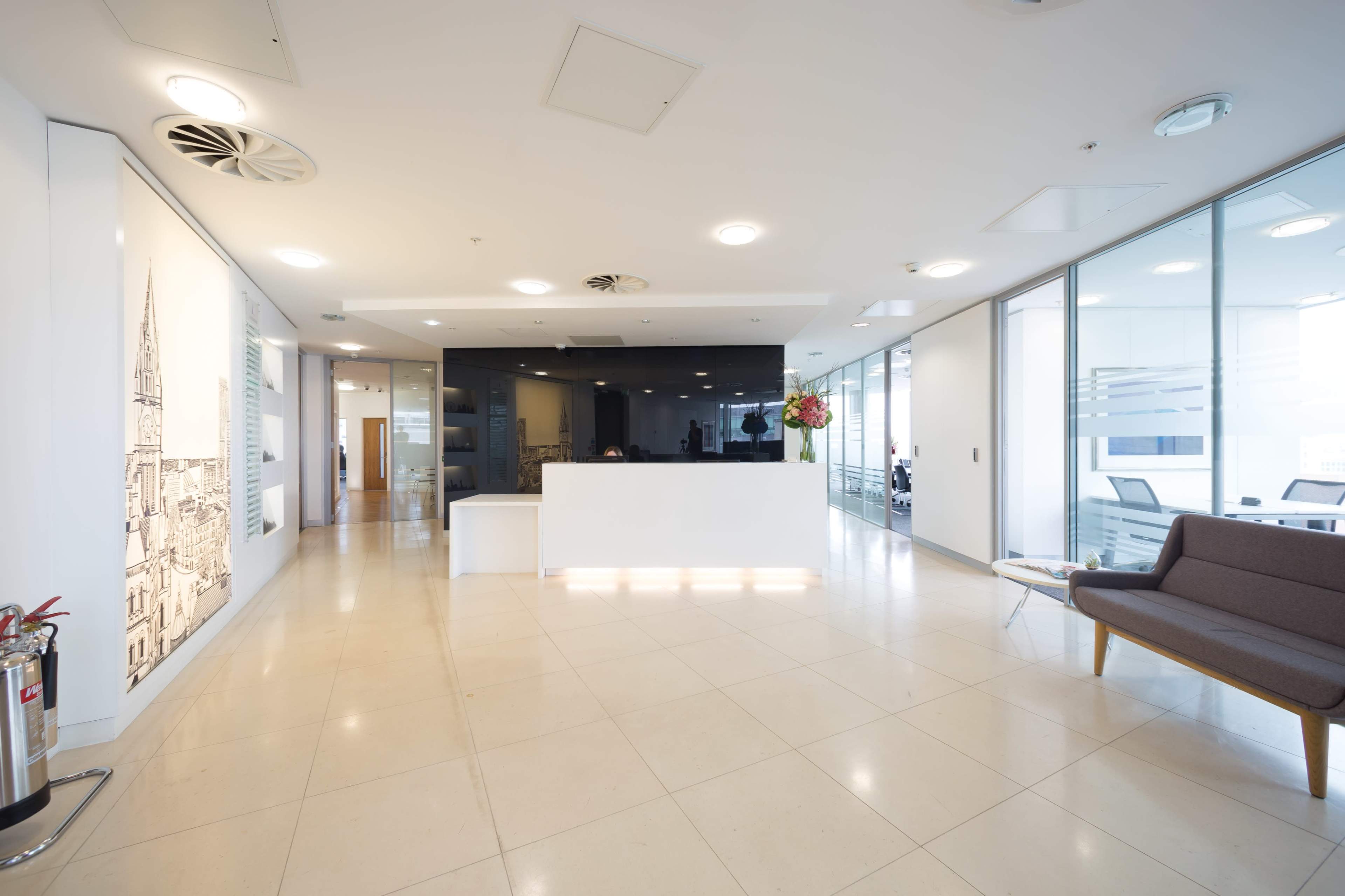 A modern office reception area with white walls, a dark reception desk, and a seating area featuring a gray couch.