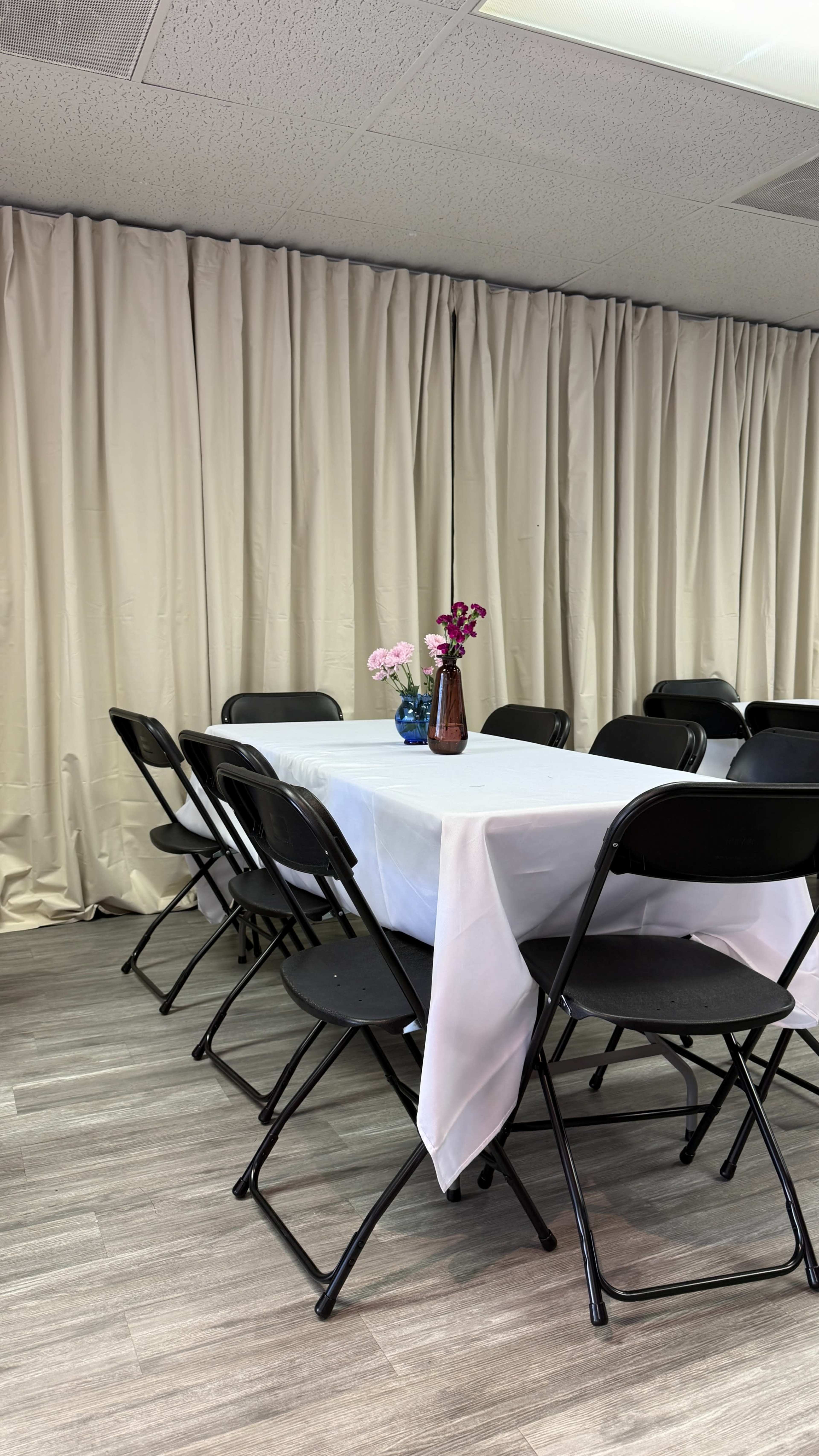 A long table covered with a white tablecloth is set in a room, surrounded by black folding chairs, and adorned with a small vase of flowers.