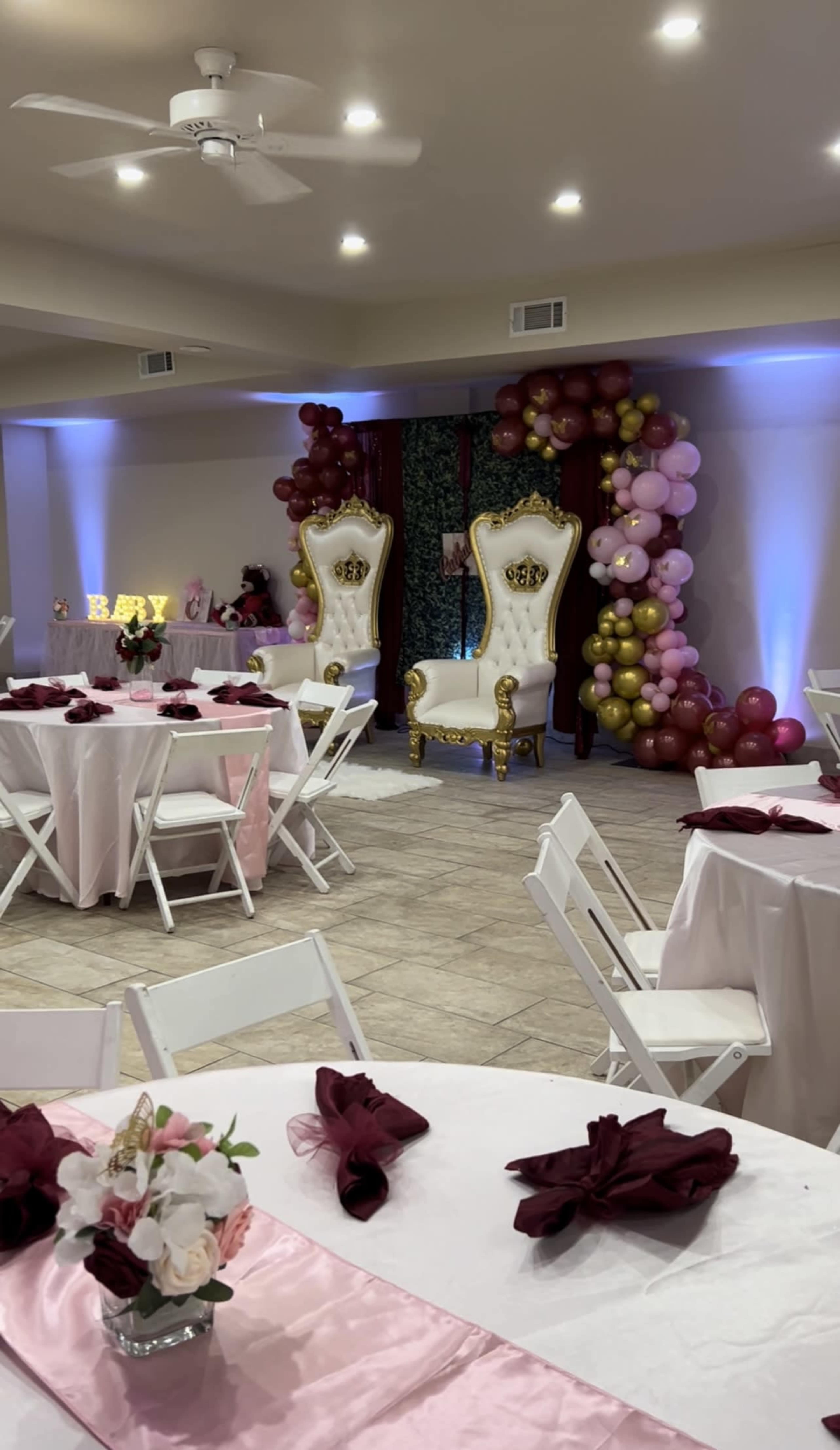 The scene features a decorated event space with white tables, maroon napkins, and two golden thrones surrounded by balloon arrangements in various colors.