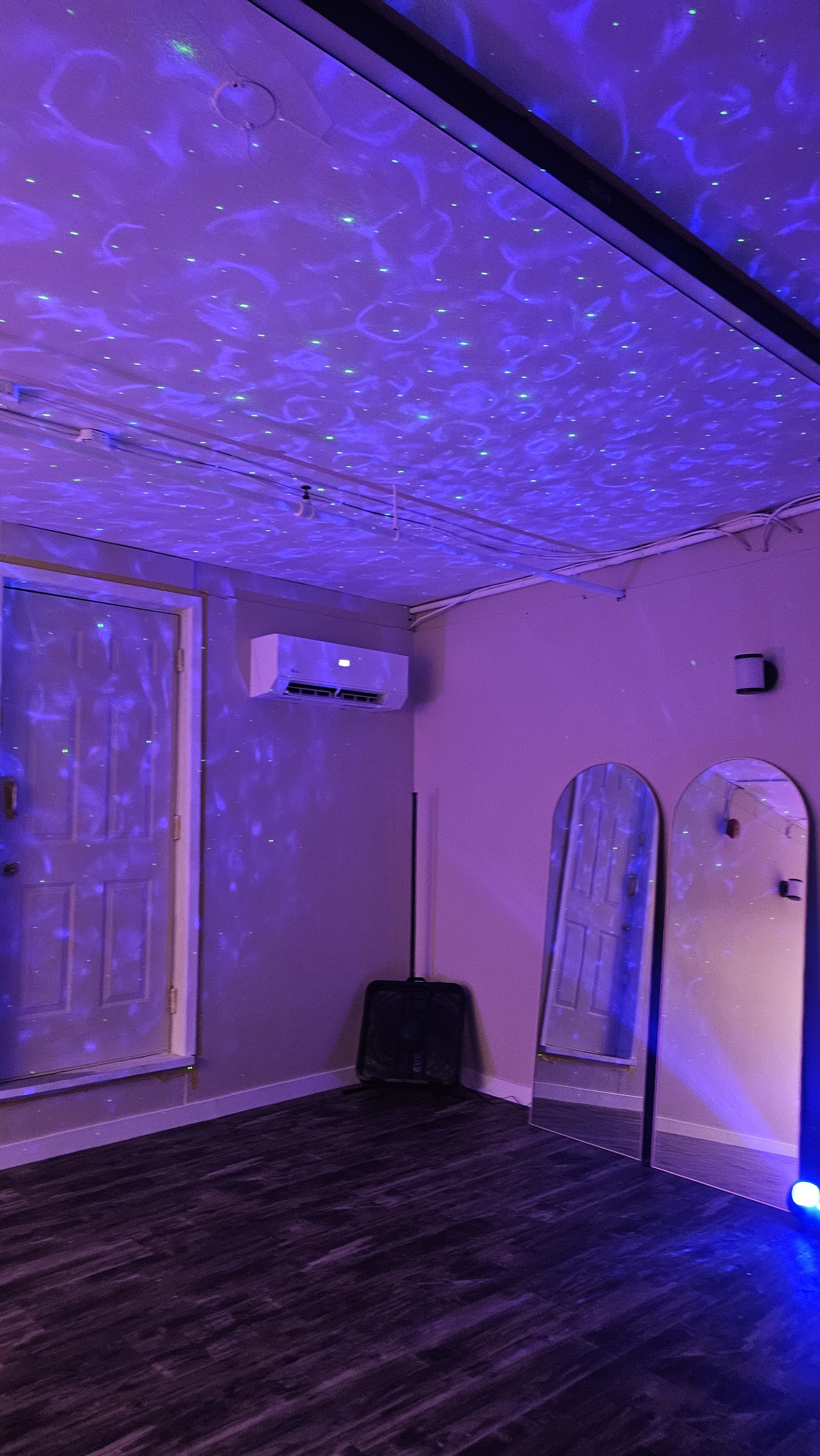 The image shows a room with a textured ceiling illuminated by blue and purple lights, featuring a door, mirrors, and a wall-mounted air conditioning unit.