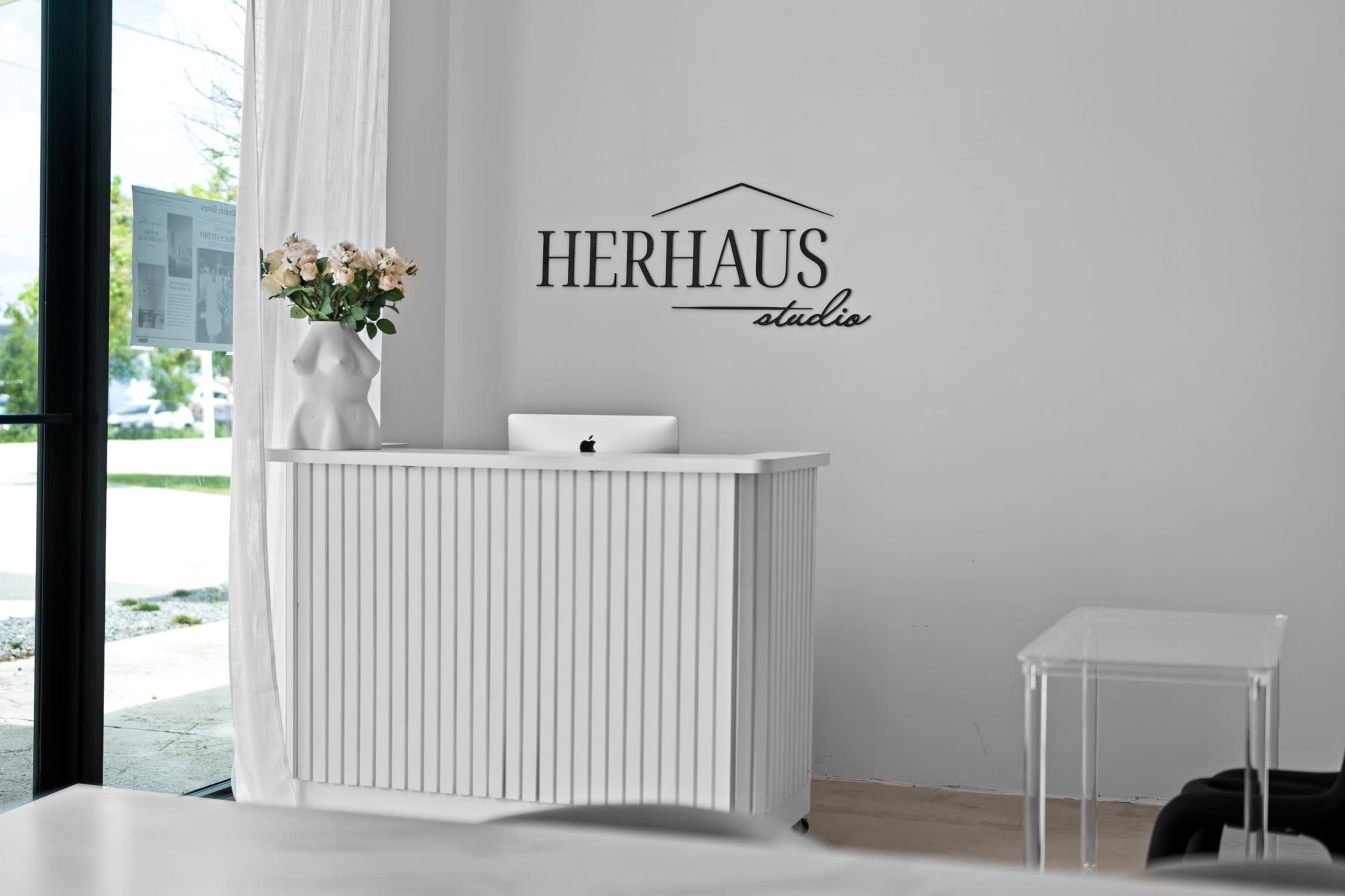 The image shows a modern reception area with a white ribbed counter, a computer on it, a vase of flowers, and a wall featuring the name "HERHAUS studio."