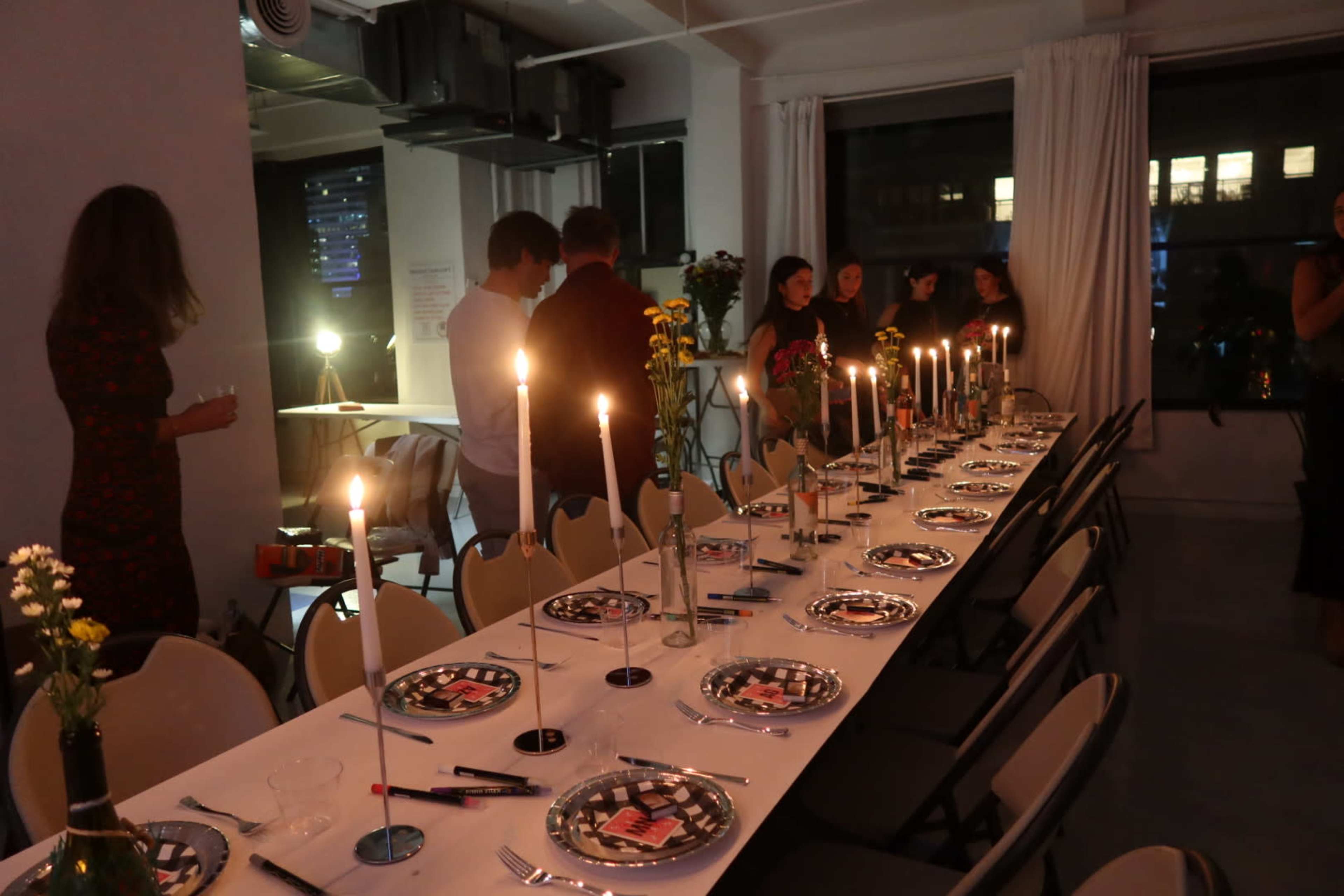 A long dining table is set with plates and candles, while guests in semi-darkness gather around in a festive setting.