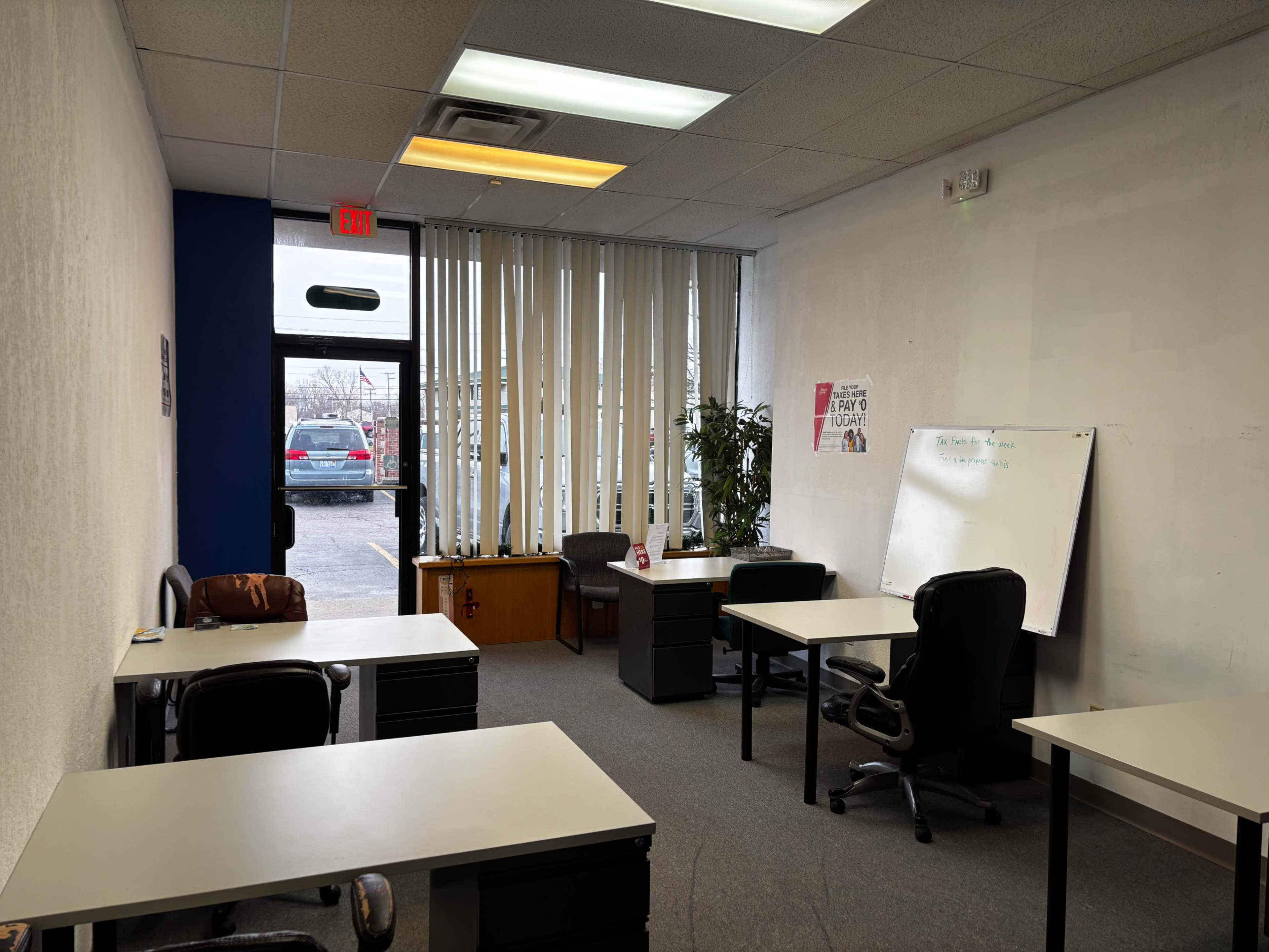 The image shows a sparsely furnished office space with several desks, chairs, and a whiteboard, illuminated by natural light from a large window.