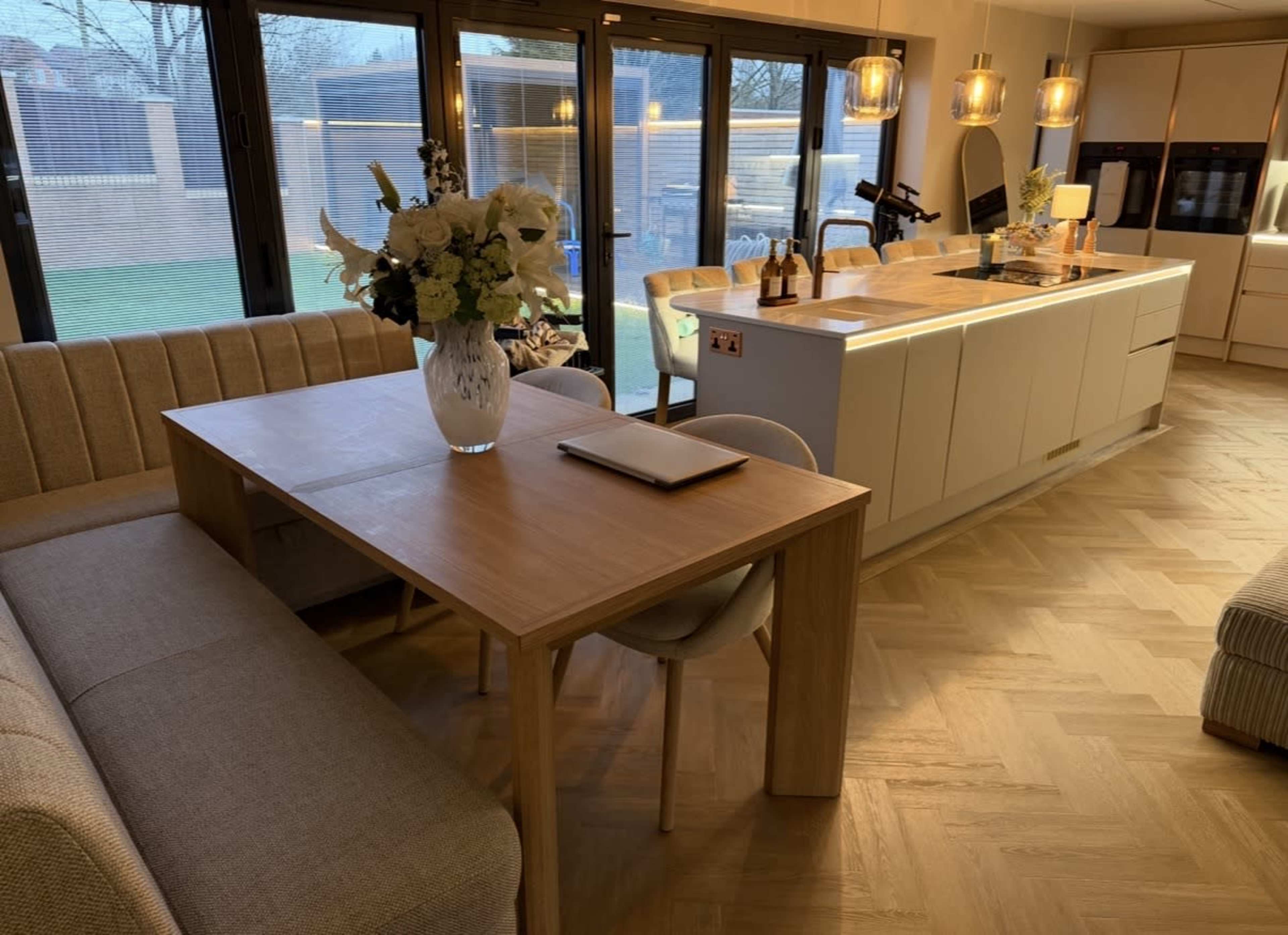 The image shows a modern kitchen and dining area featuring a wooden table with a vase of flowers, a beige couch, and a kitchen island with pendant lights and appliances.
