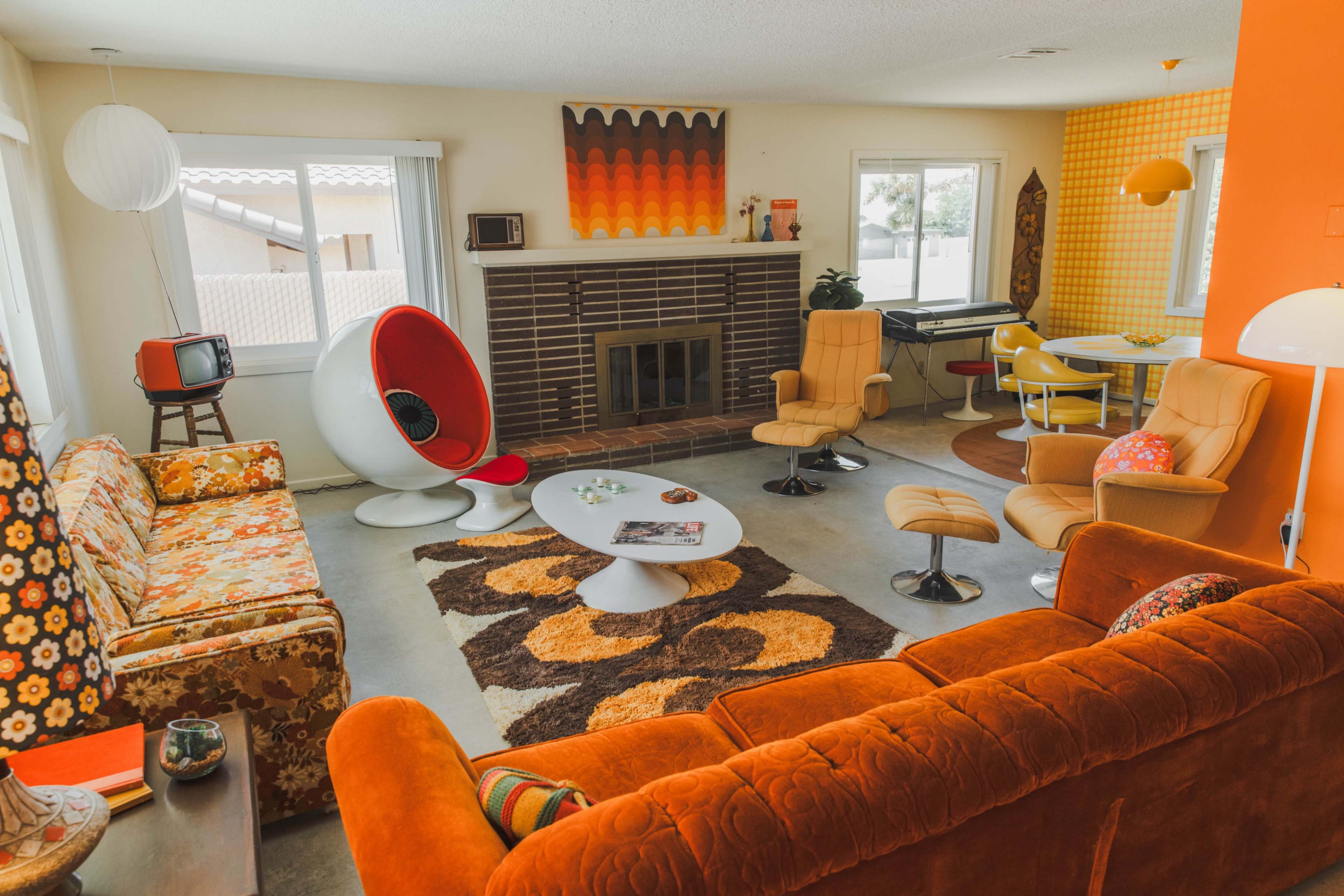 A retro living room with orange and floral-patterned furniture, a circular coffee table, and a fireplace, complemented by a dining area featuring a small round table and colorful chairs.